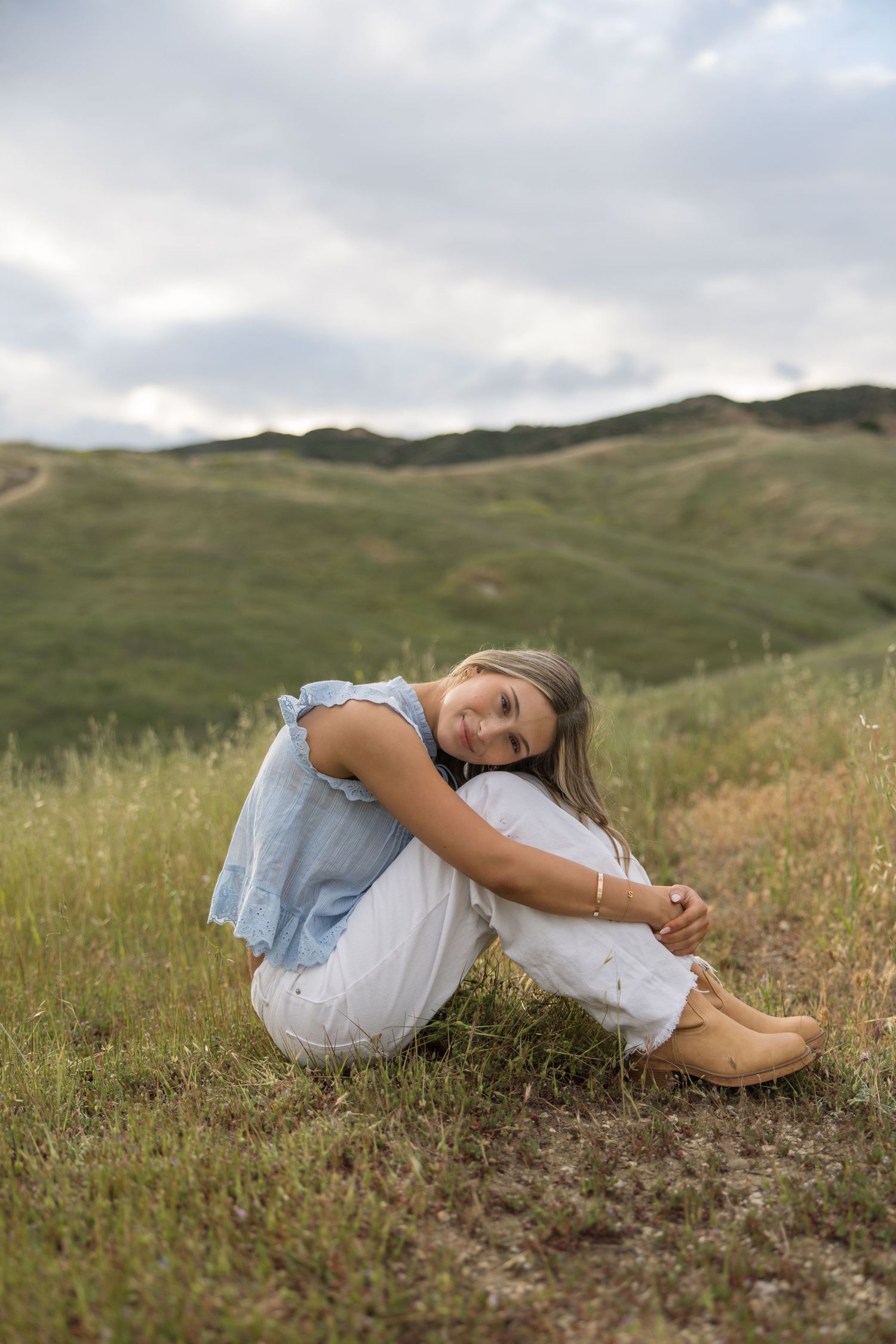 Young woman in blue top and white pants sitting on grass with green hills in the background.