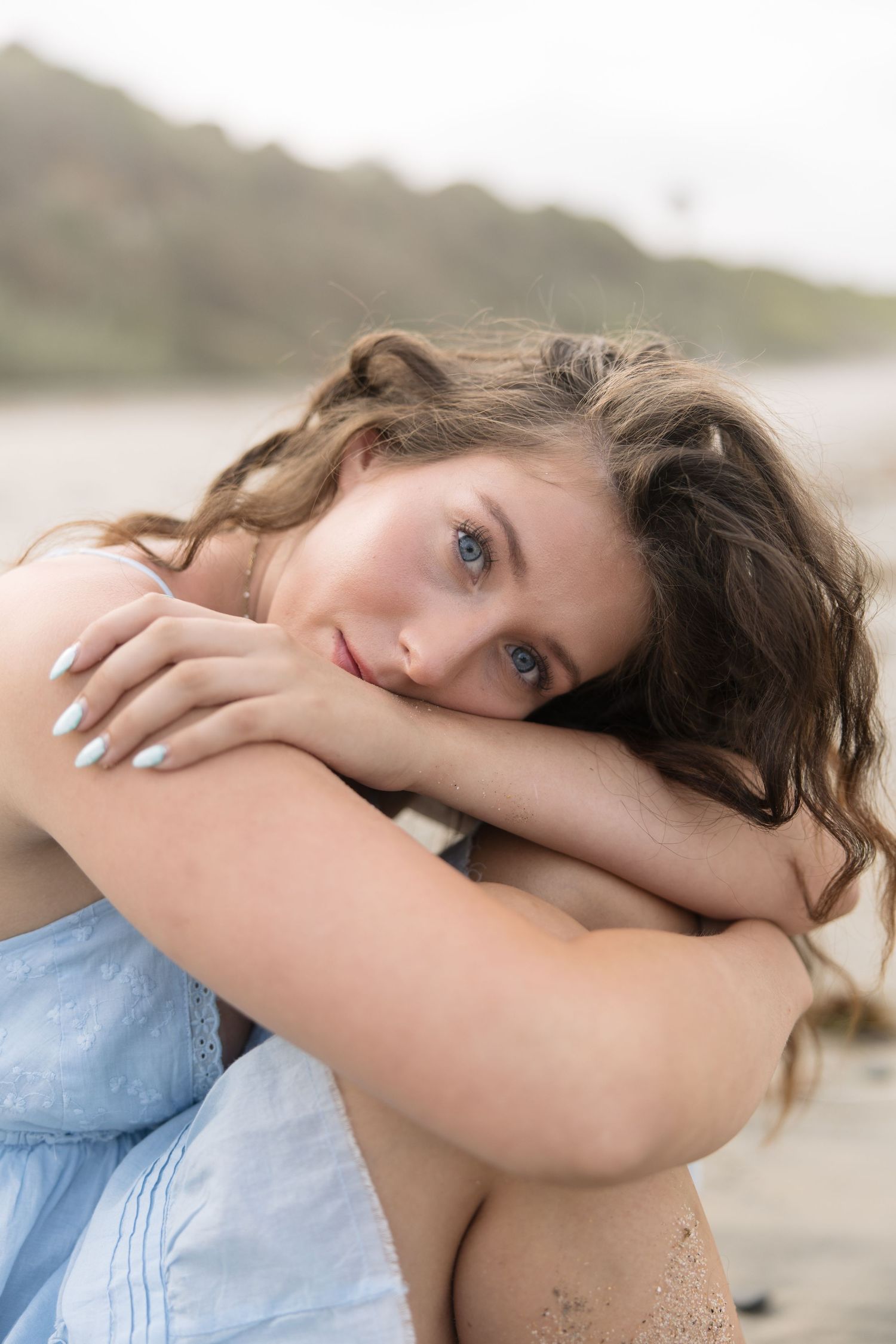 Young woman with blue eyes and wavy brown hair resting her chin on her knees at a beach.