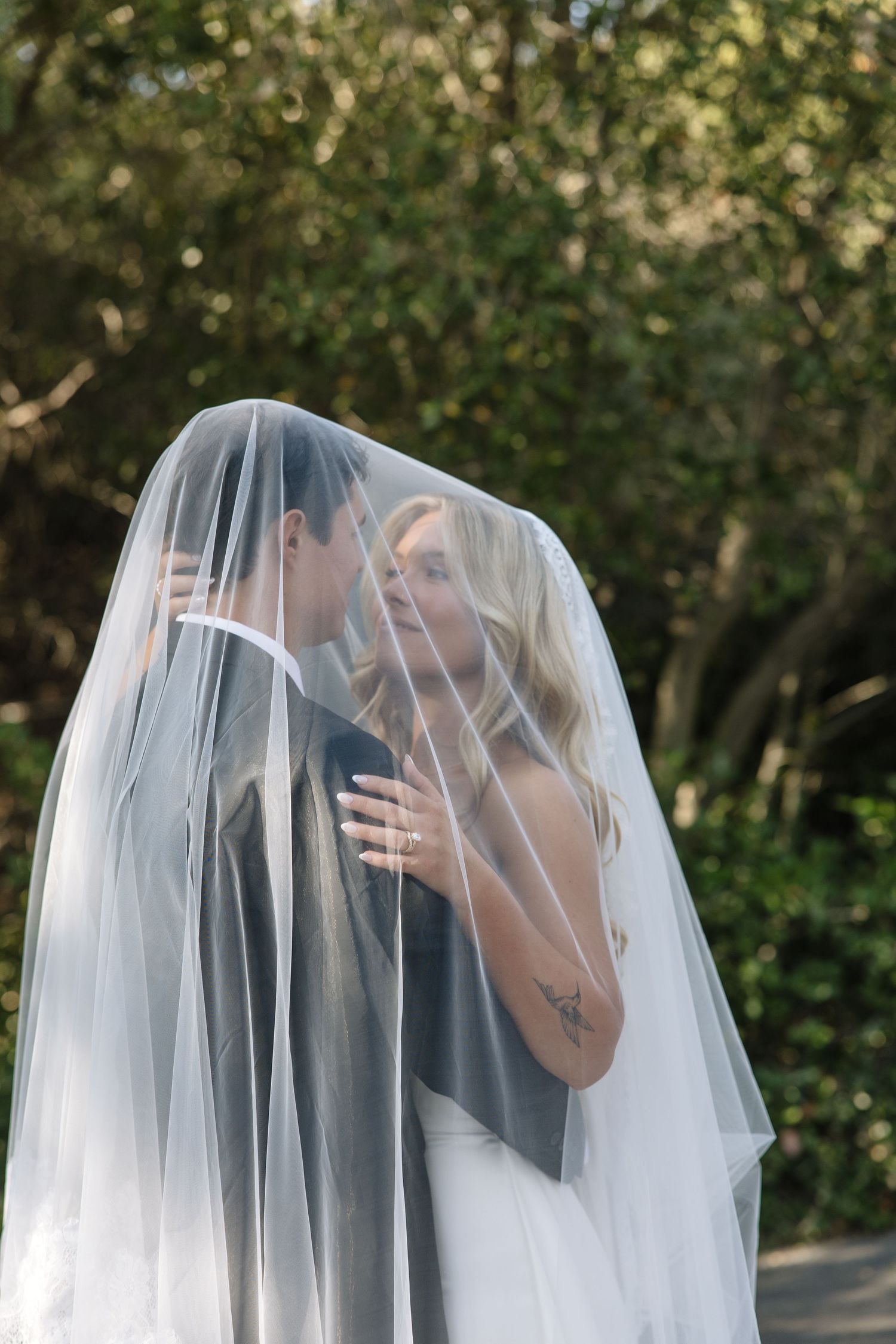 Bride and groom share a romantic kiss under a flowing white wedding veil outdoors surrounded by lush green trees.