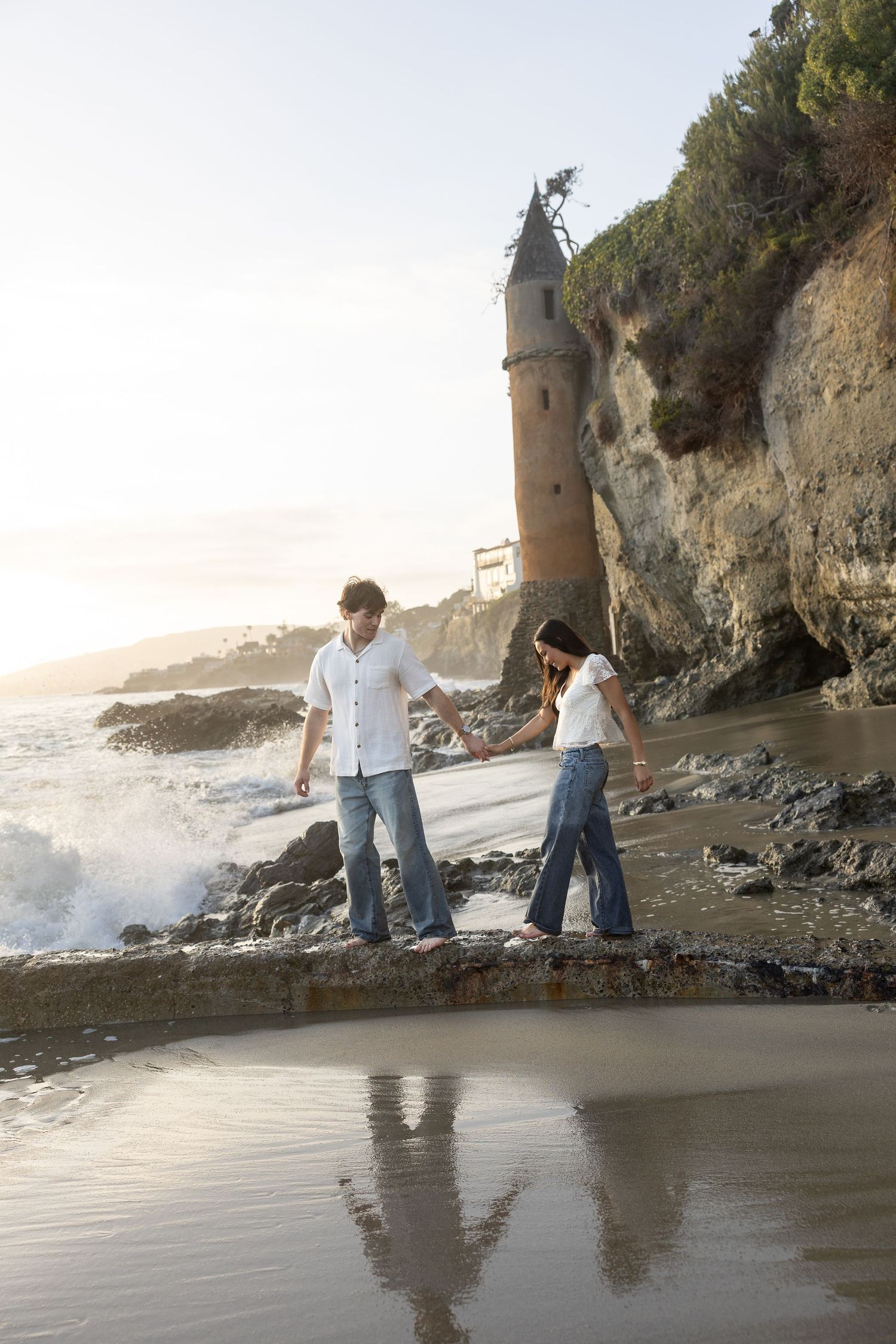 Couple holding hands on rocky beach at sunset with historic stone tower on coastal cliff in background.