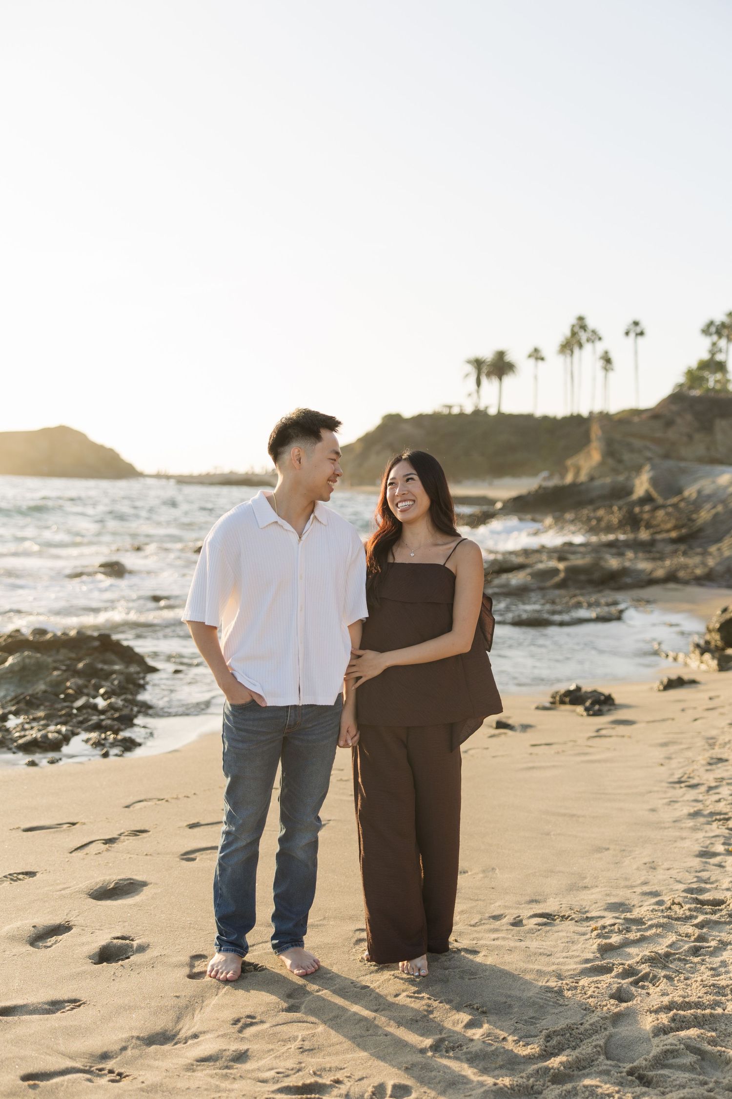 Smiling couple walking barefoot along a sandy beach at sunset with rocky cliffs, ocean waves, and palm trees in the background.