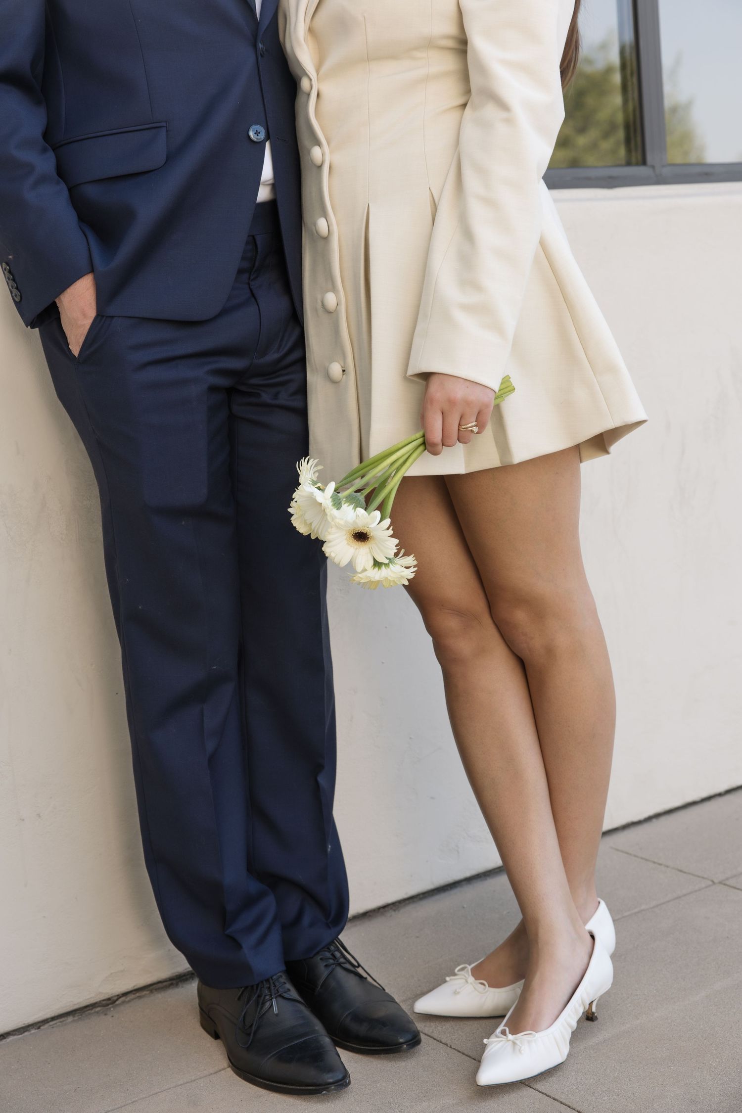 Couple standing against a wall, she in a cream button-front dress and white heels holding white daisies, he in a navy suit.