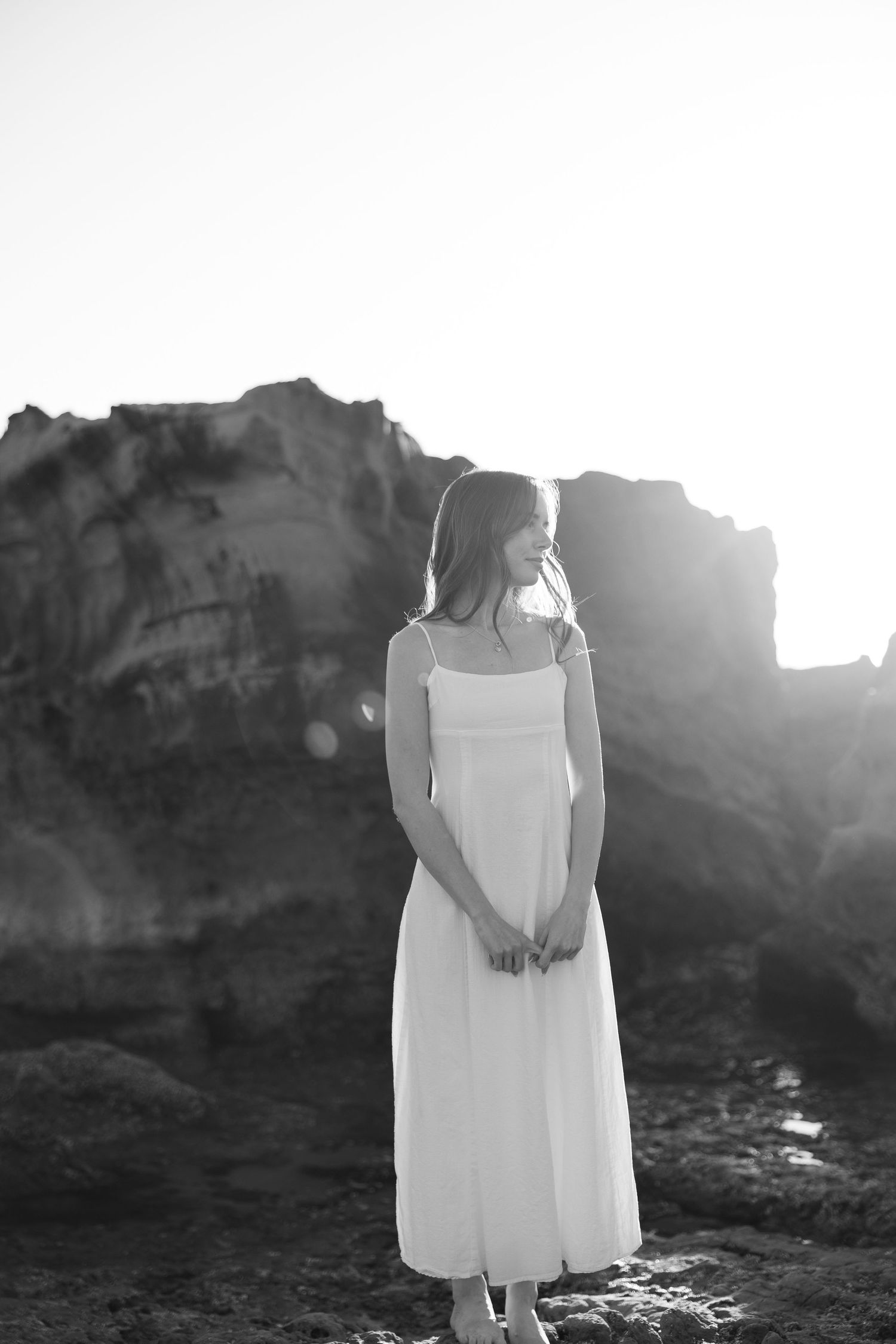 Black and white photo of a woman in a white sundress standing on rocky coastal cliffs with sunlight behind her.