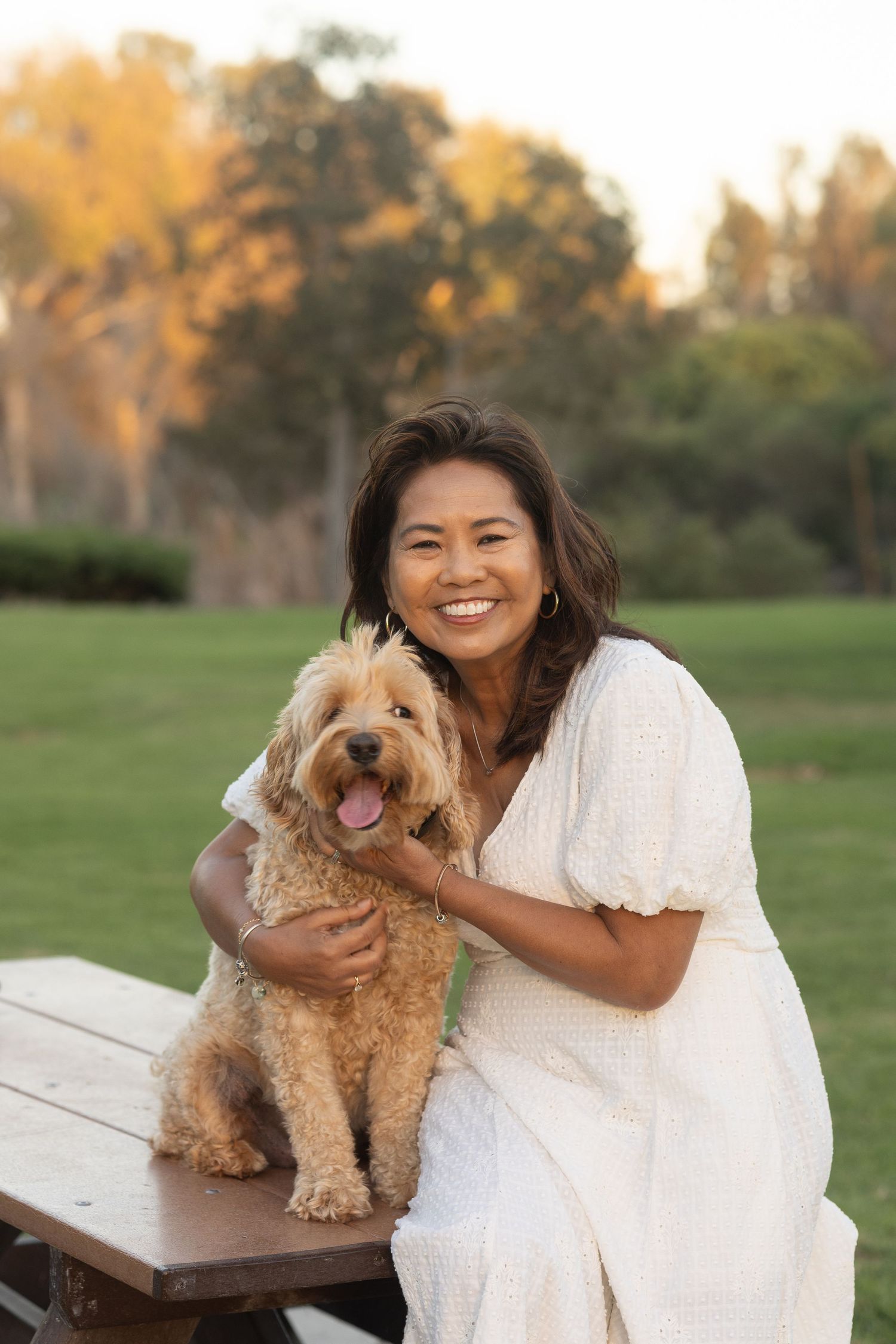 Person in white dress hugging a happy goldendoodle dog in a park setting.