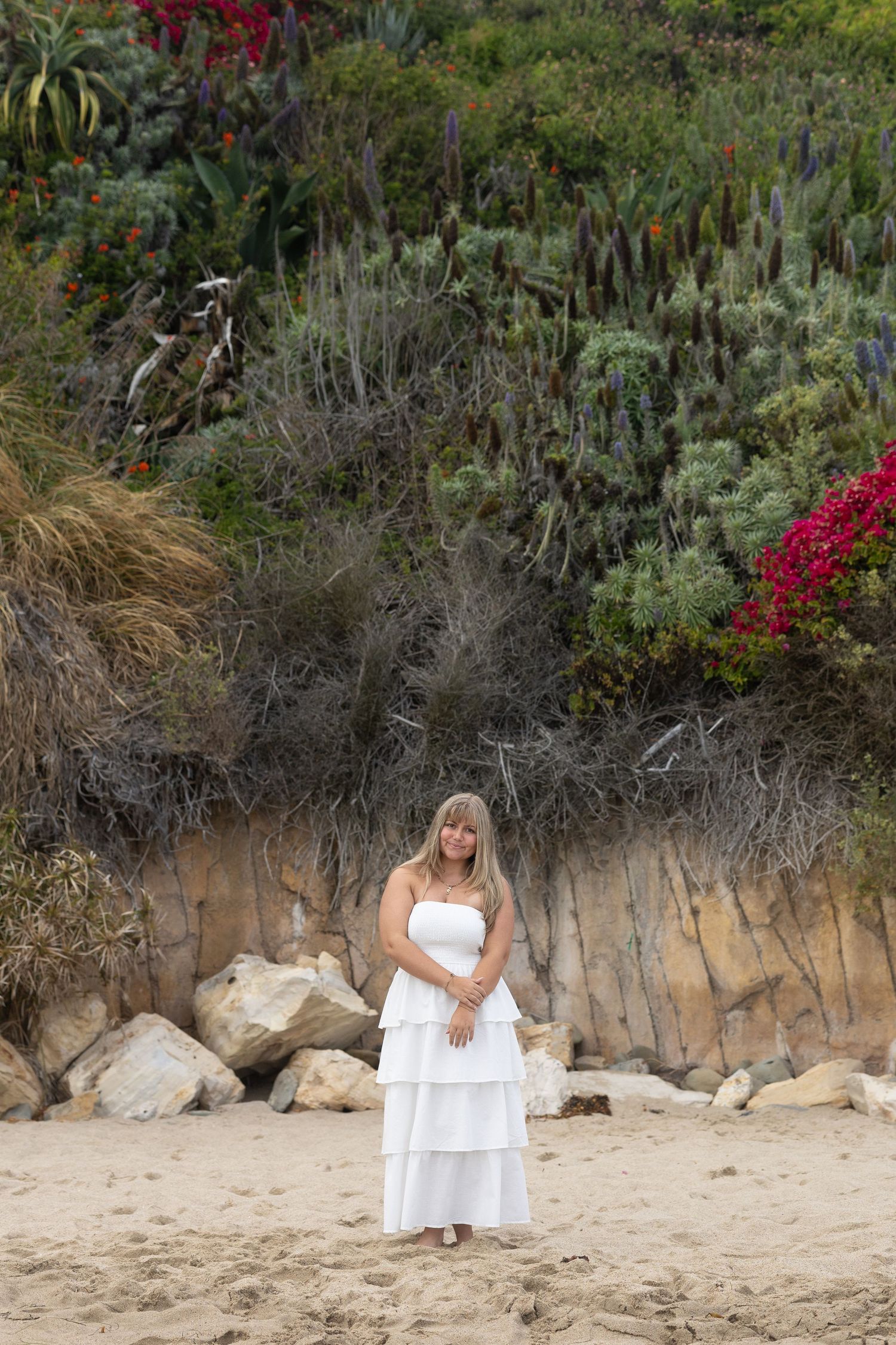 Beachside portrait session with white dress against stone wall and flowering plants.
