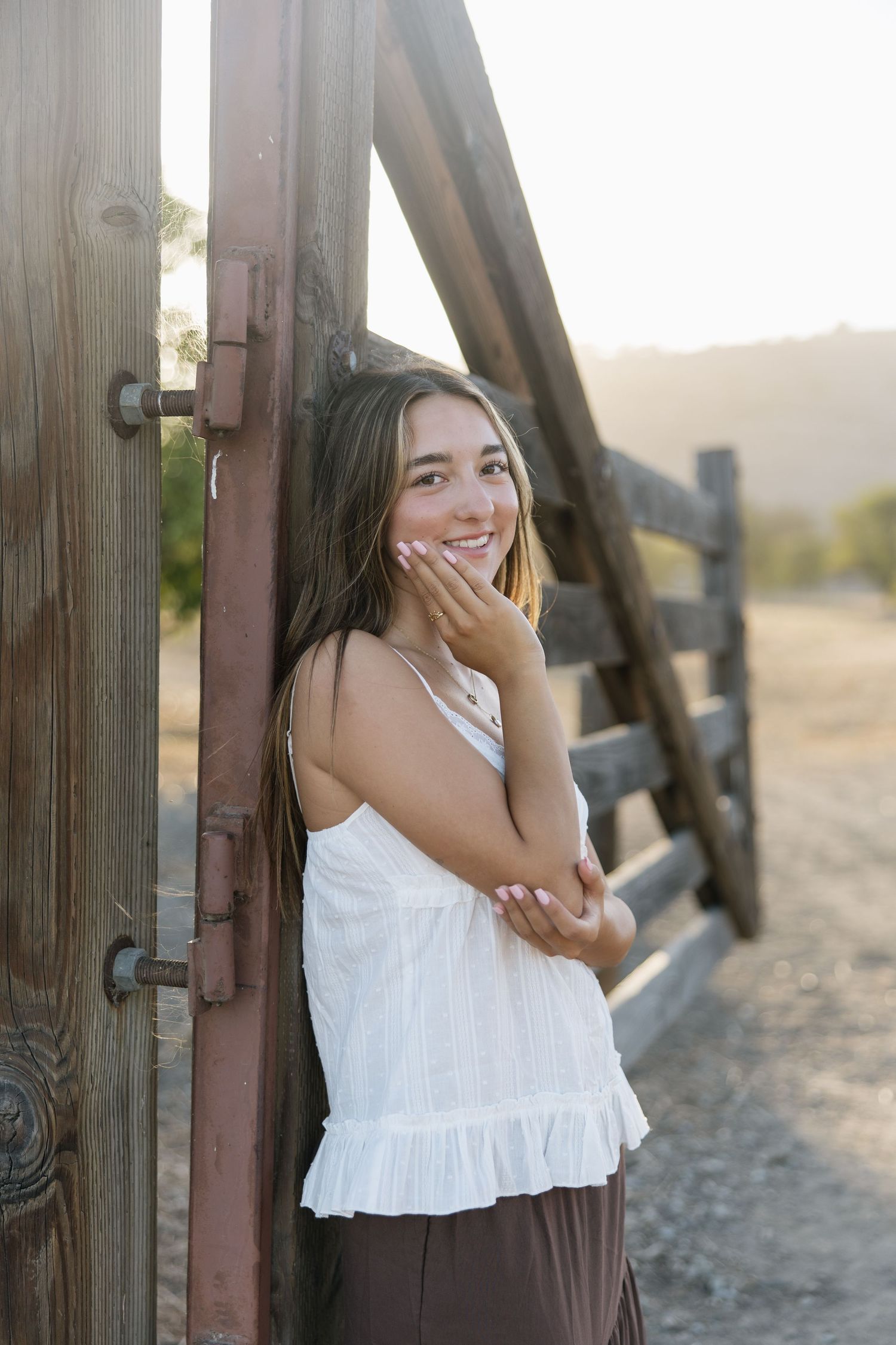 Smiling teenage girl in white top leaning against rustic wooden farm gate at golden hour.