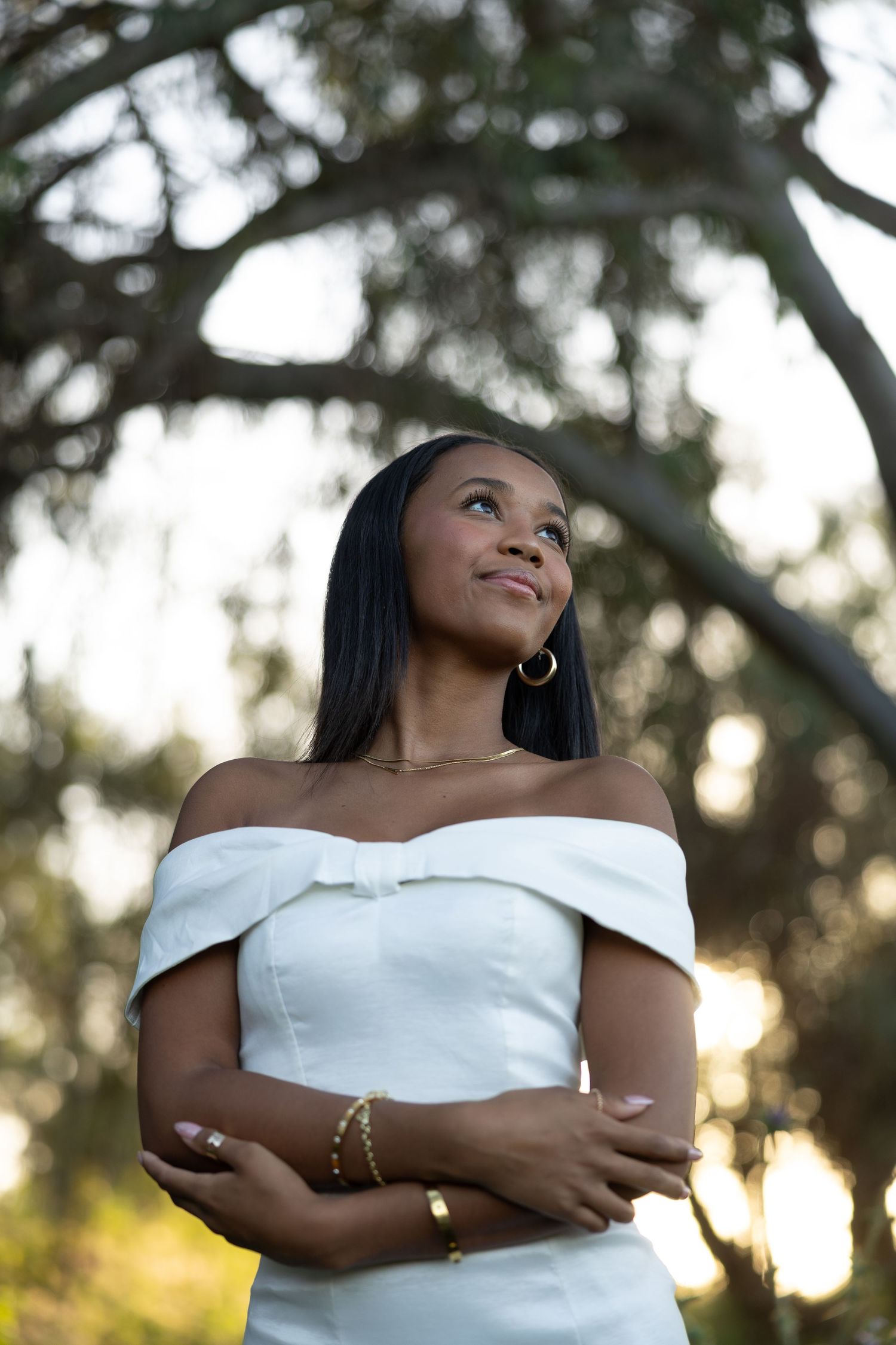 A young Black woman in a white off-shoulder dress with a bow stands outdoors, arms crossed, smiling upward near trees.