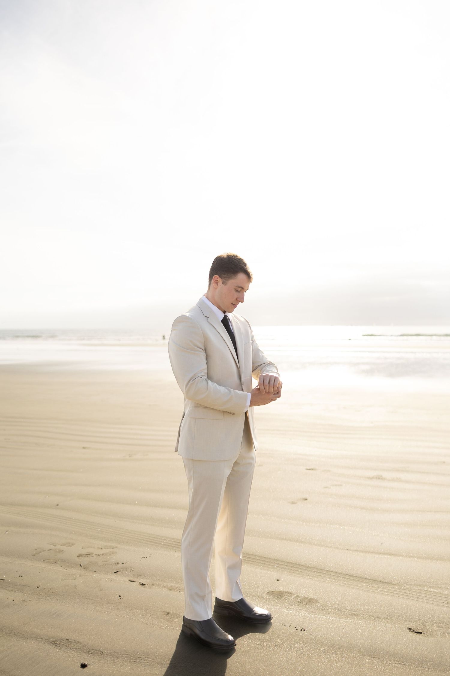 A person in a beige suit checks their watch while standing on a sandy beach at sunset.