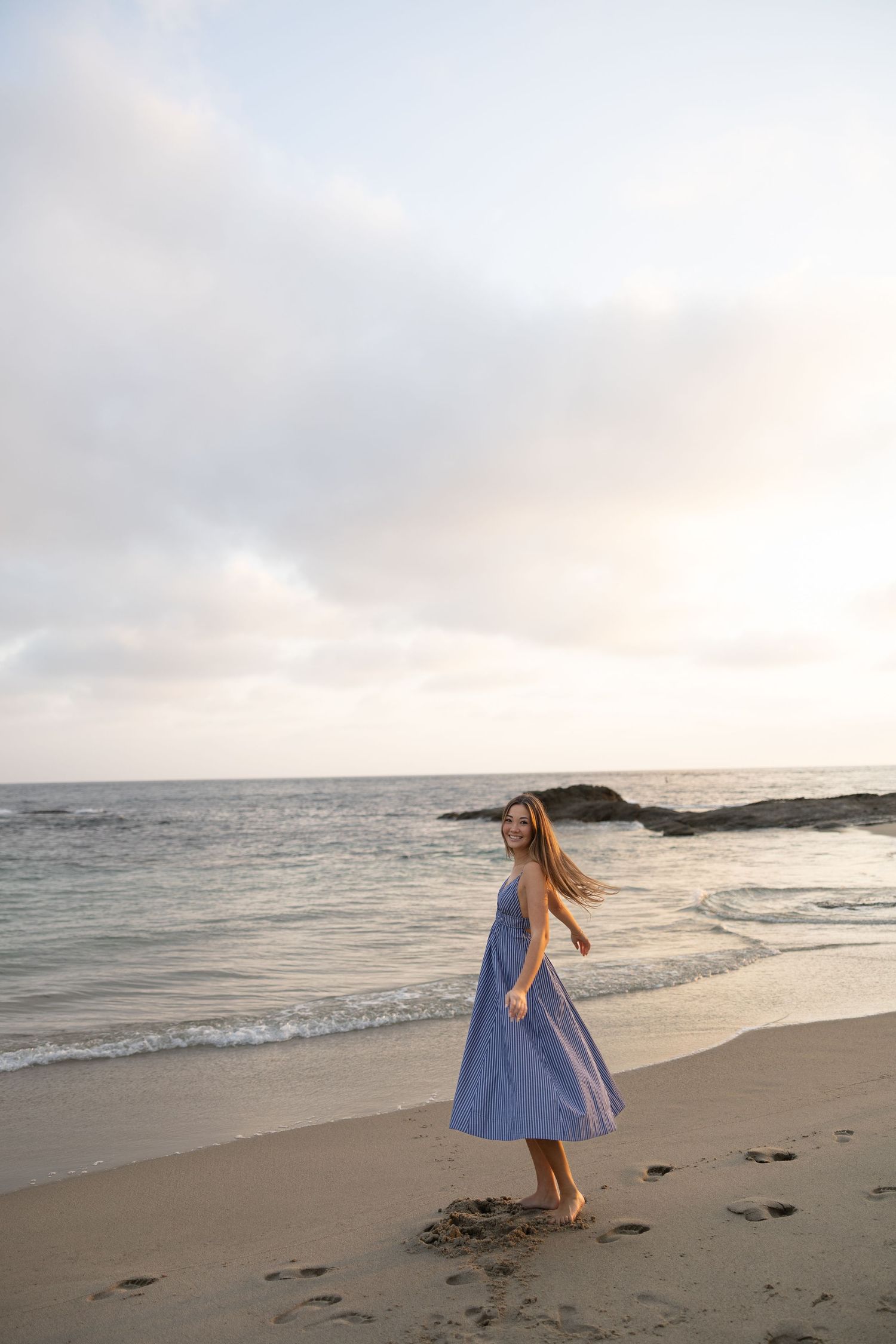 A young woman in a blue striped dress walks barefoot on a sandy beach at sunset with ocean waves behind her.
