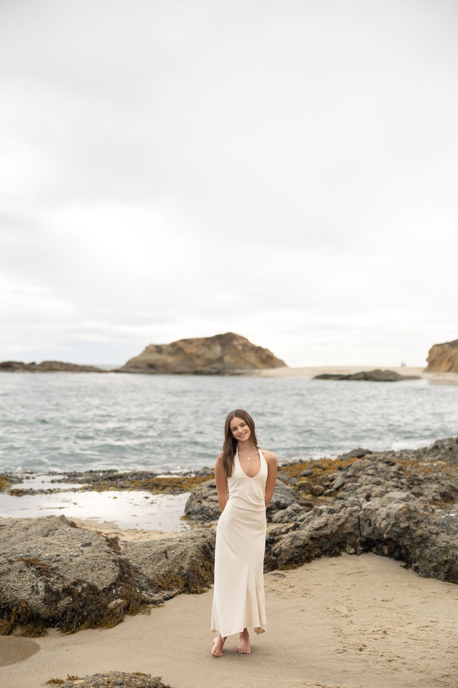 Woman in white maxi dress standing on rocky beach with ocean and coastal rock formations in background.