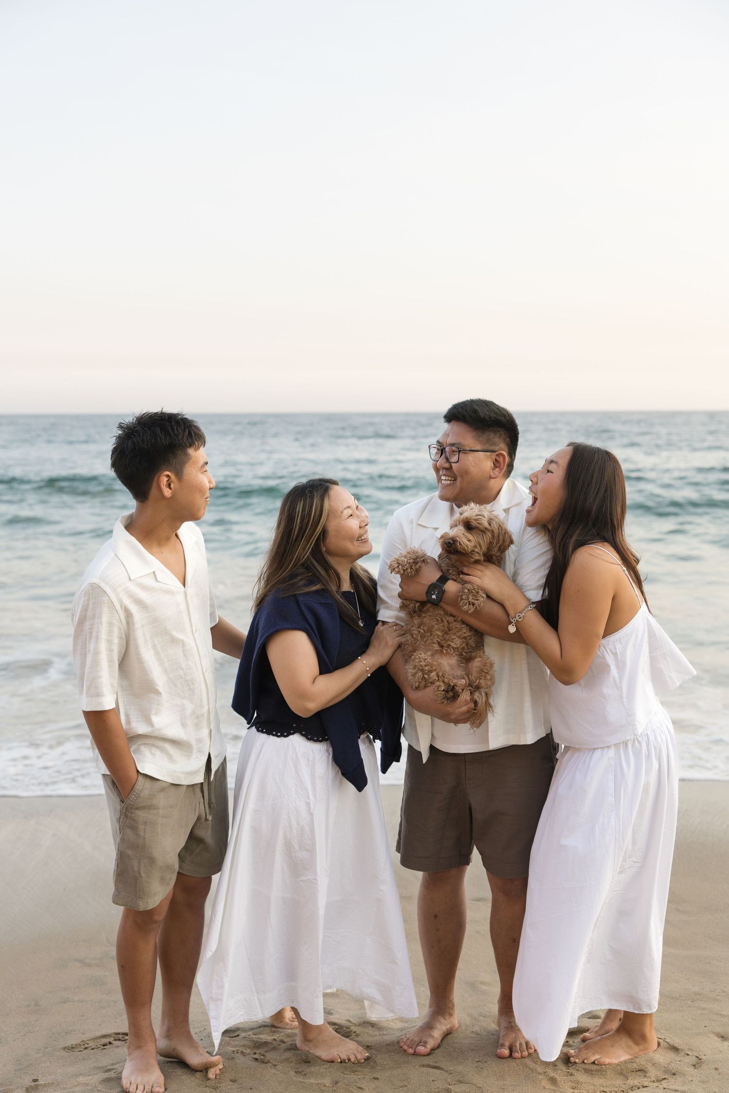 Four people and a small brown dog enjoy a fun moment together on a sandy beach with ocean waves in the background.