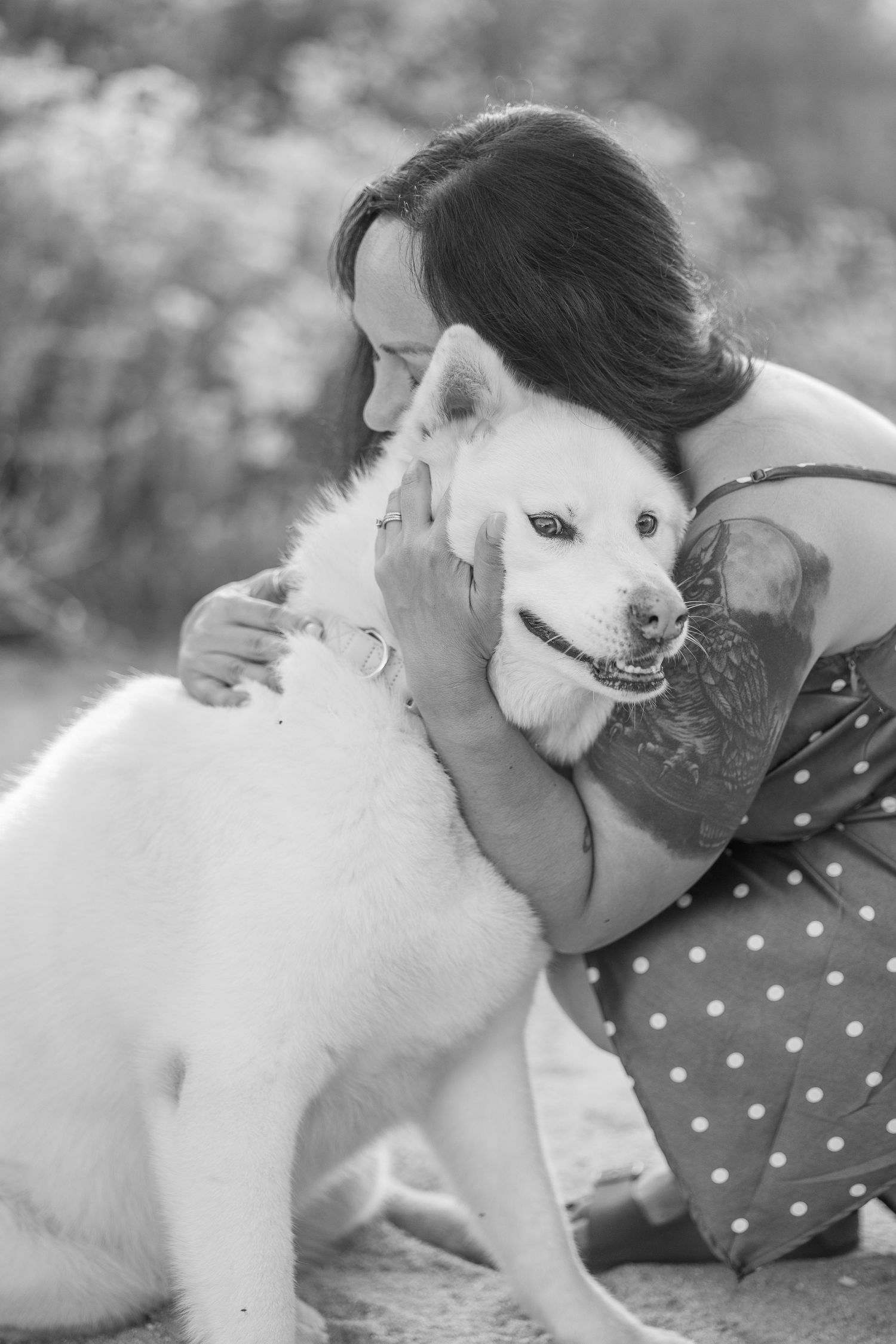 Black and white portrait of person embracing a white dog in a polka dot dress.