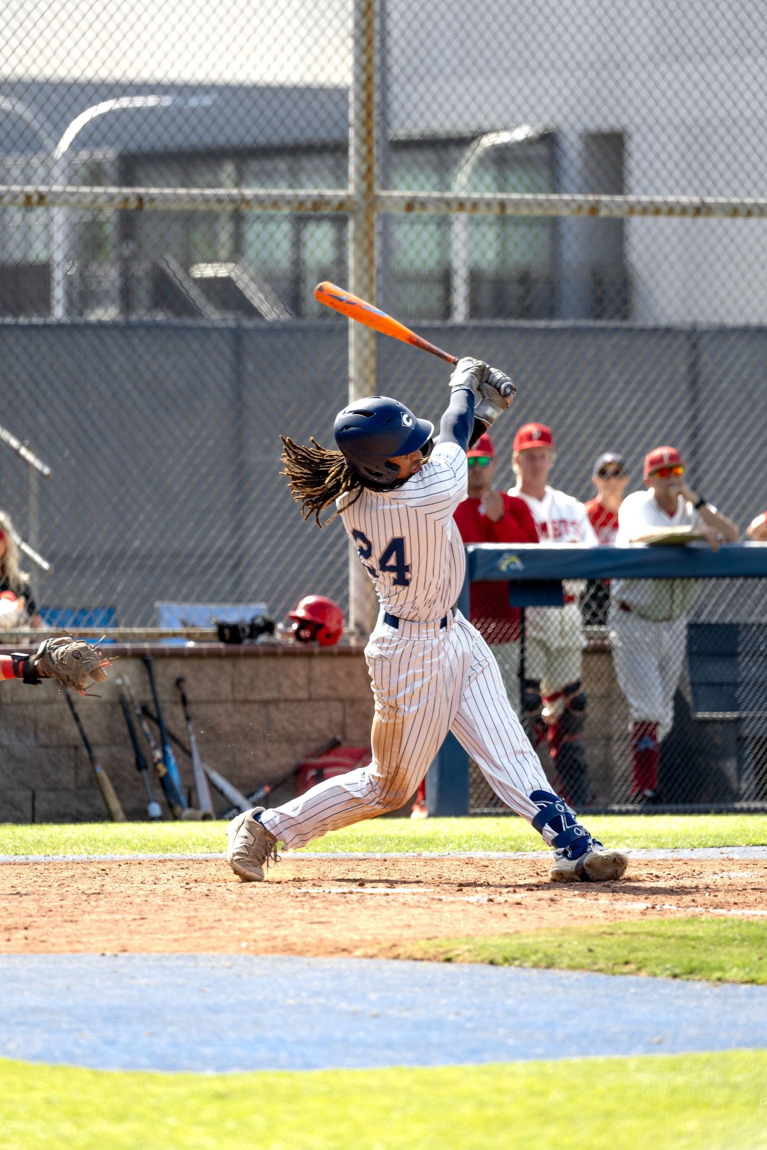 Baseball player #24 in white pinstripe uniform swings orange bat during a game with fans watching in background.