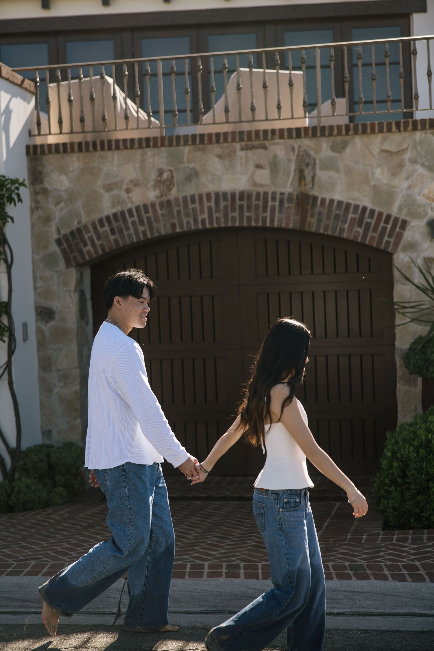 Young Asian couple holding hands walking in front of a stone building with arched garage doors.