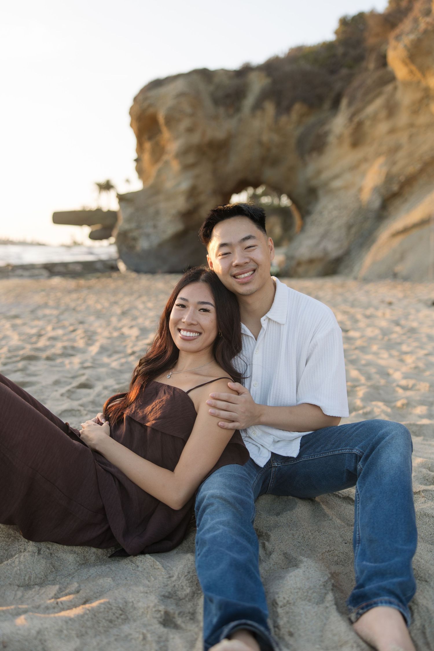 Smiling couple sitting close together on a sandy beach at sunset with a rocky sea arch in the background.