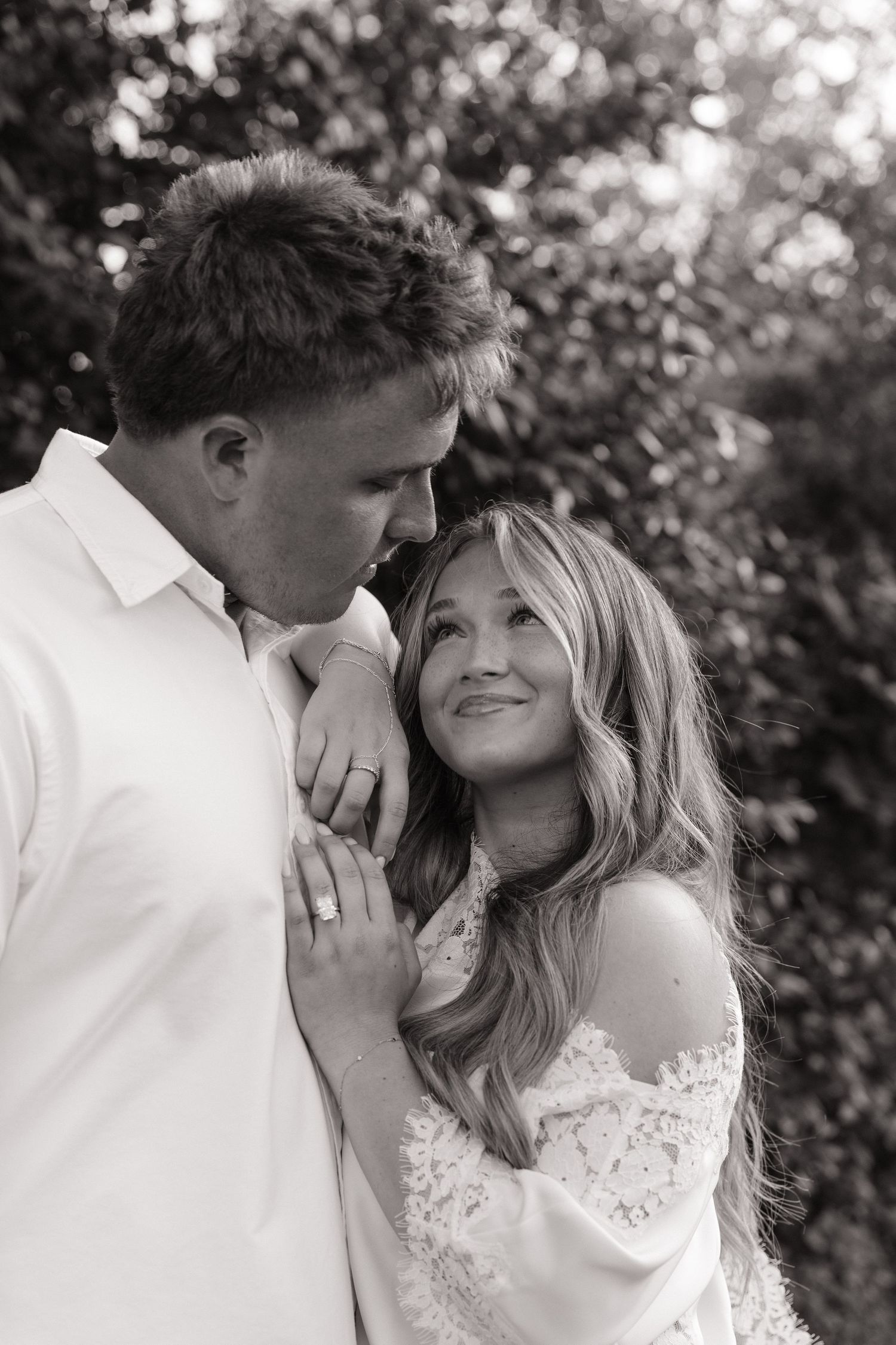 Black and white photo of a smiling bride leaning on groom's shoulder outdoors with lush greenery background.