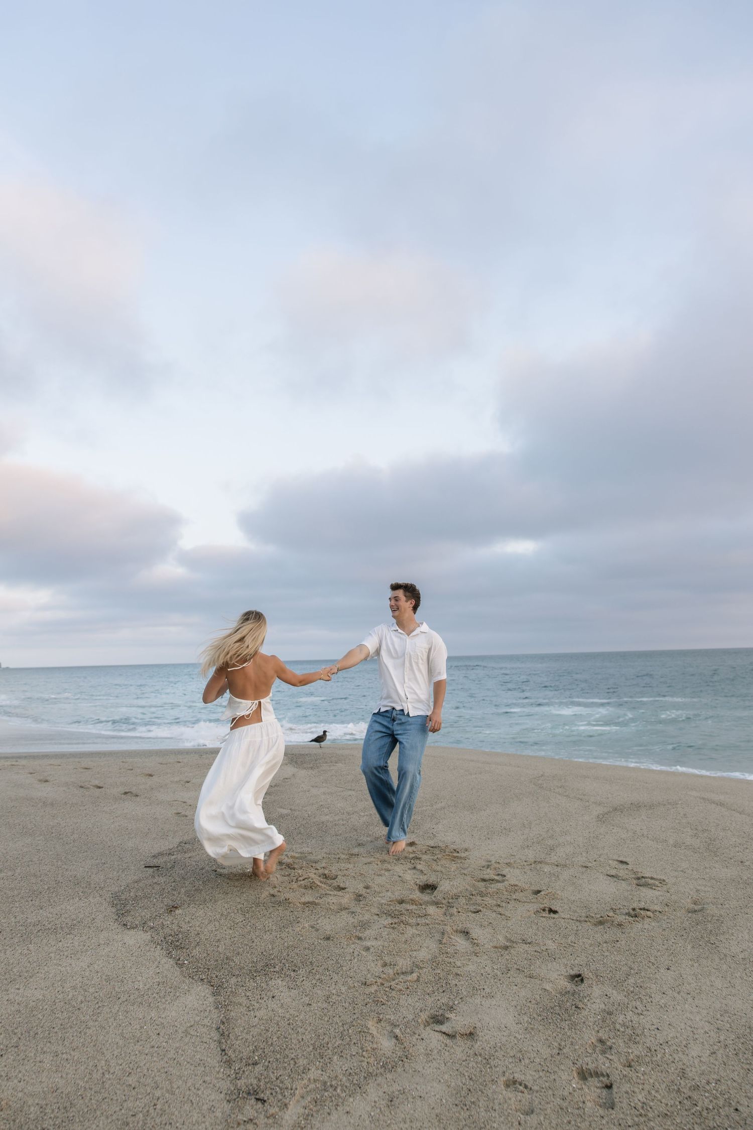 Couple dancing barefoot on sandy beach at sunset, woman in white flowing dress, man in jeans and white shirt, ocean backdrop.