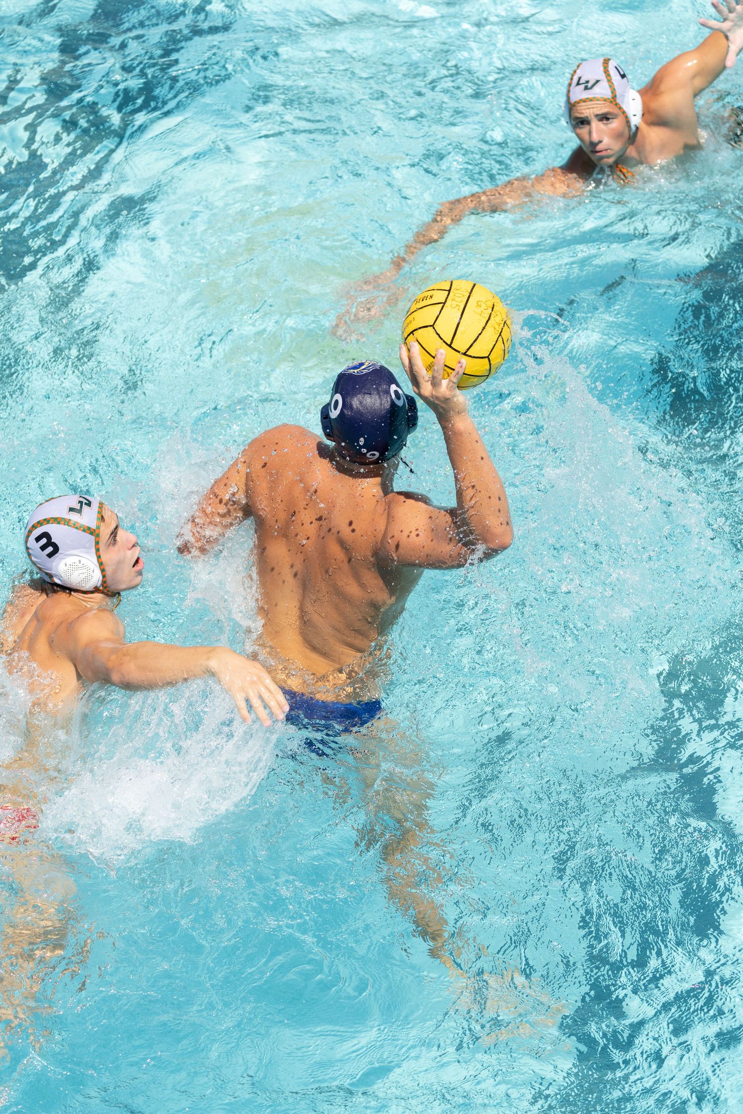 Three male water polo players compete in a pool, with one player holding a yellow ball preparing to throw.
