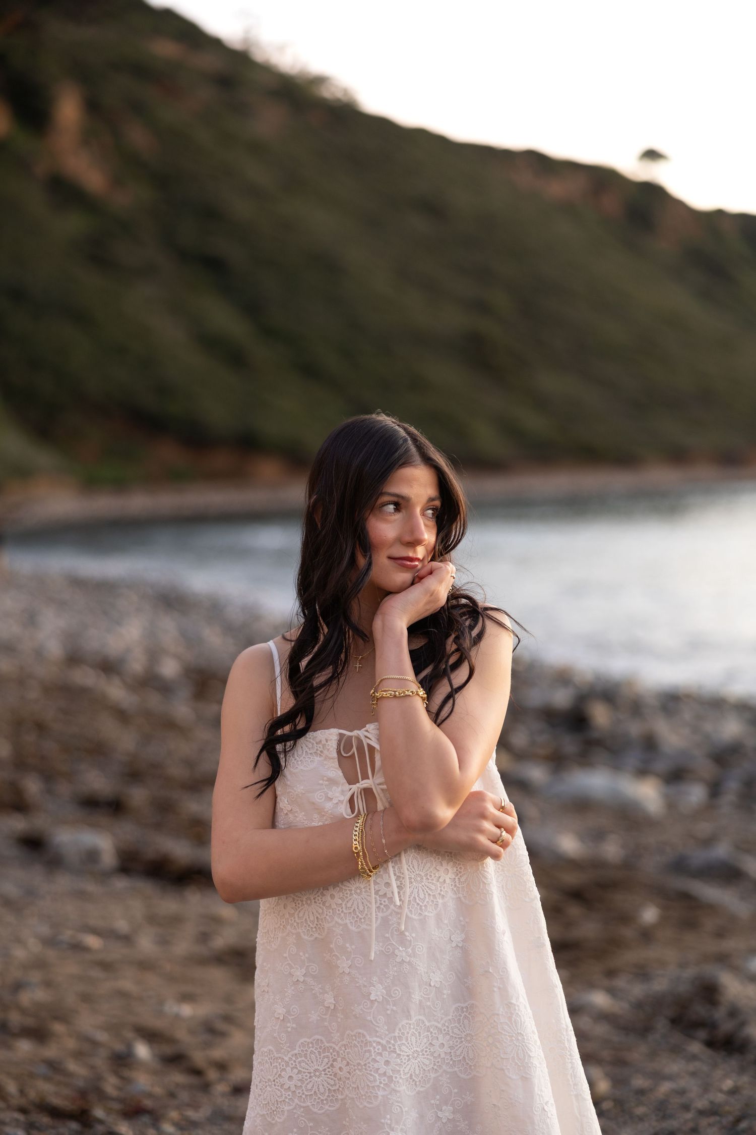 Person in a white lace dress poses on a rocky beach with cliffs and ocean in the background.