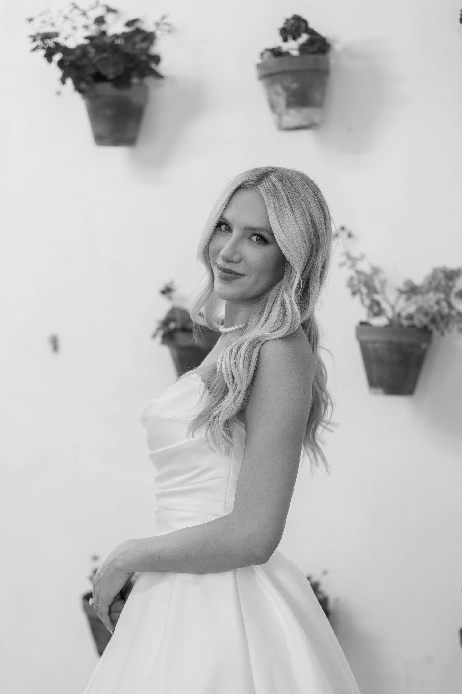 A bride in a classic white ballgown poses against a white wall with potted plants.