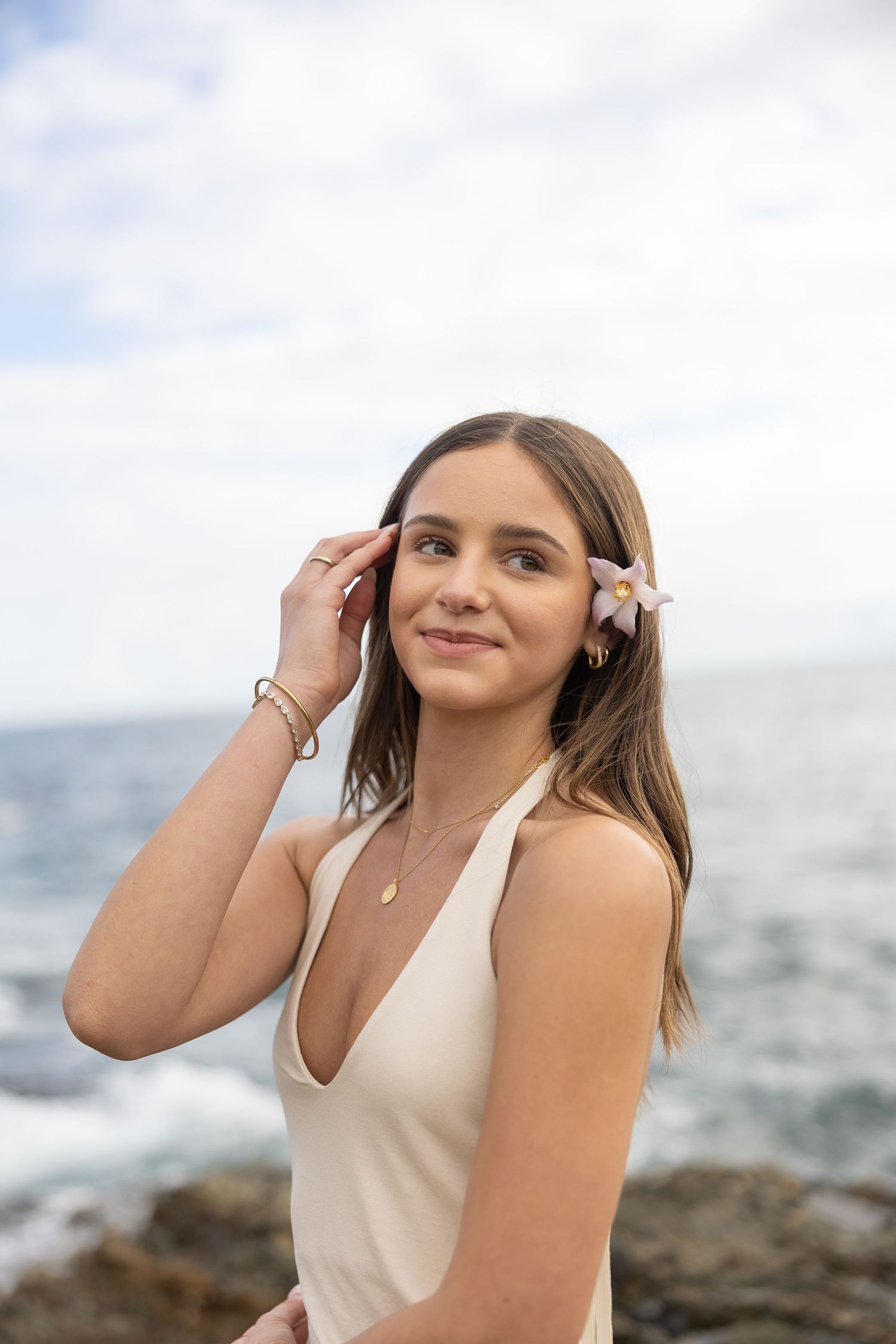Young woman in cream swimsuit with flower in hair smiling by rocky ocean shore.