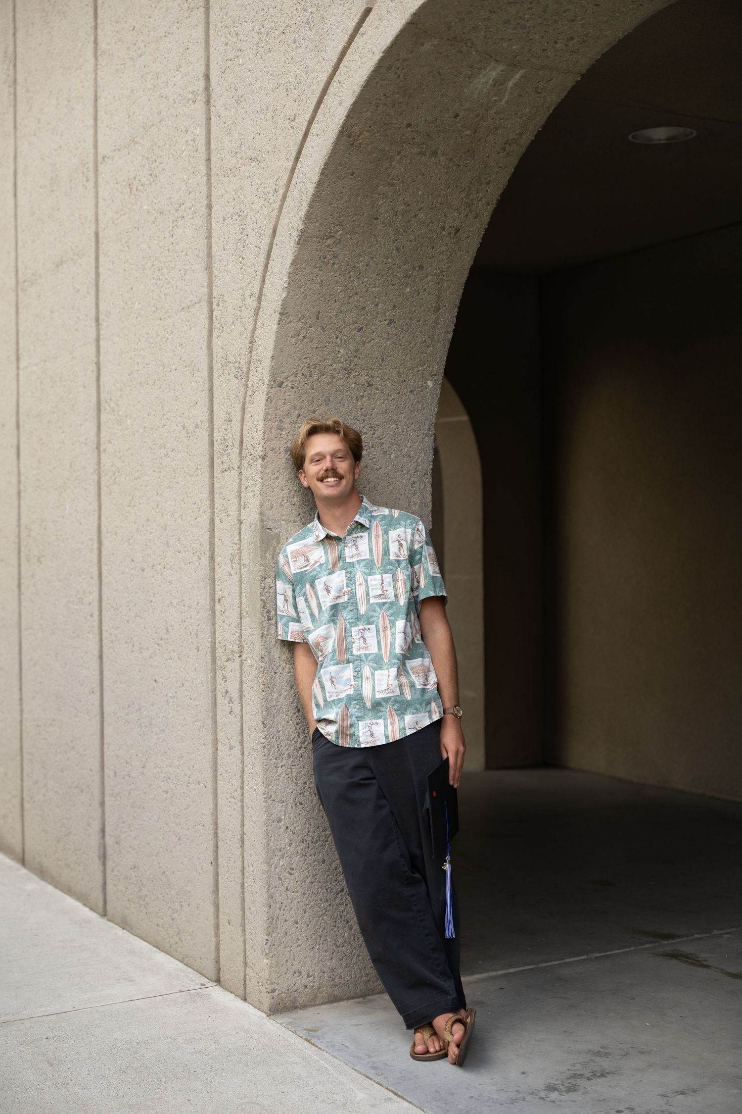 Young man with mustache leaning against stone archway, wearing floral shirt and dark pants, smiling outdoors.