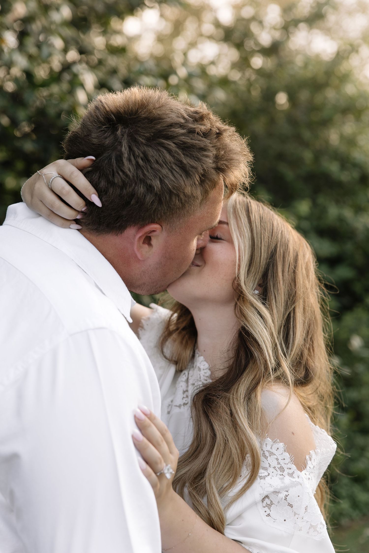 A couple in white attire shares a romantic kiss outdoors, with lush green foliage in the background.