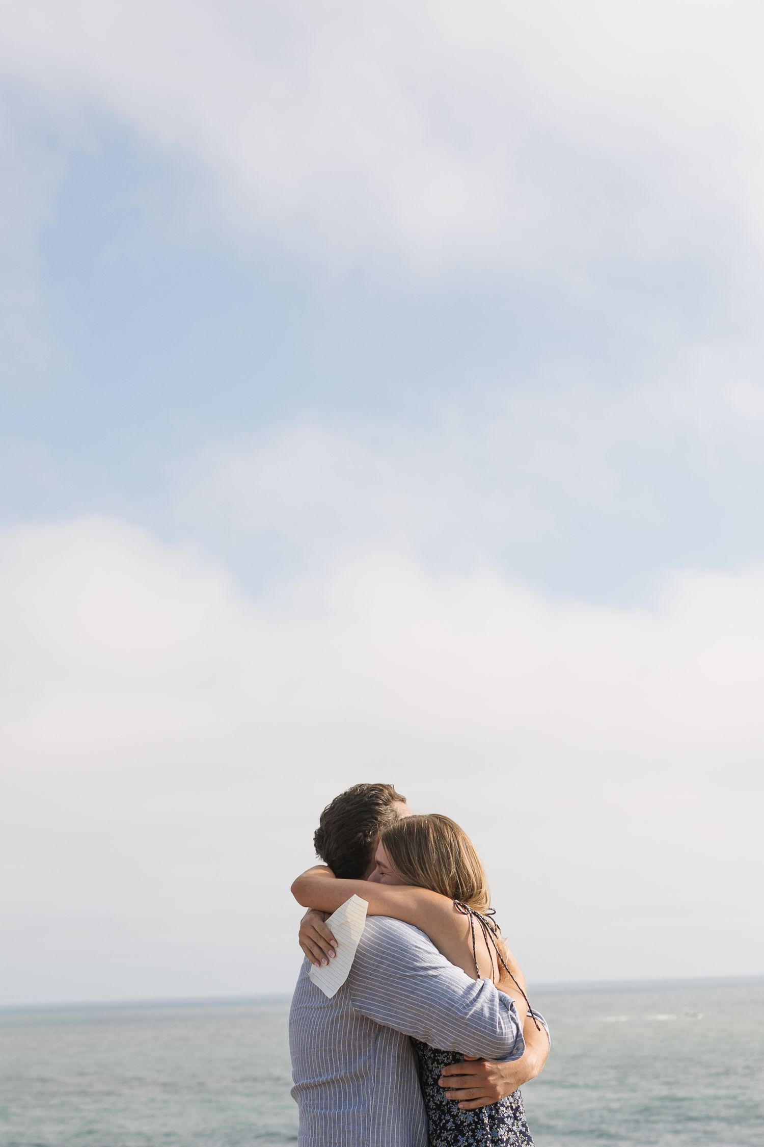 Couple embracing romantically by the ocean under a cloudy sky, sharing a tender hug with the sea in the background.