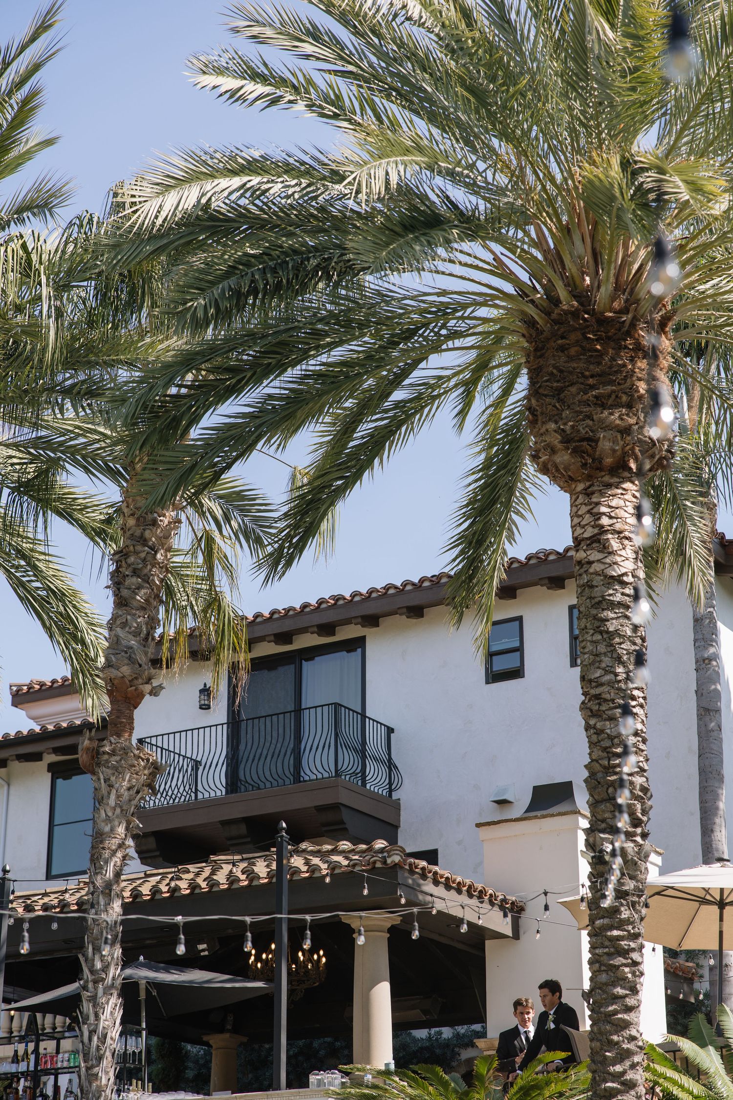Spanish-style building with palm trees, balcony, and outdoor dining area under blue sky.
