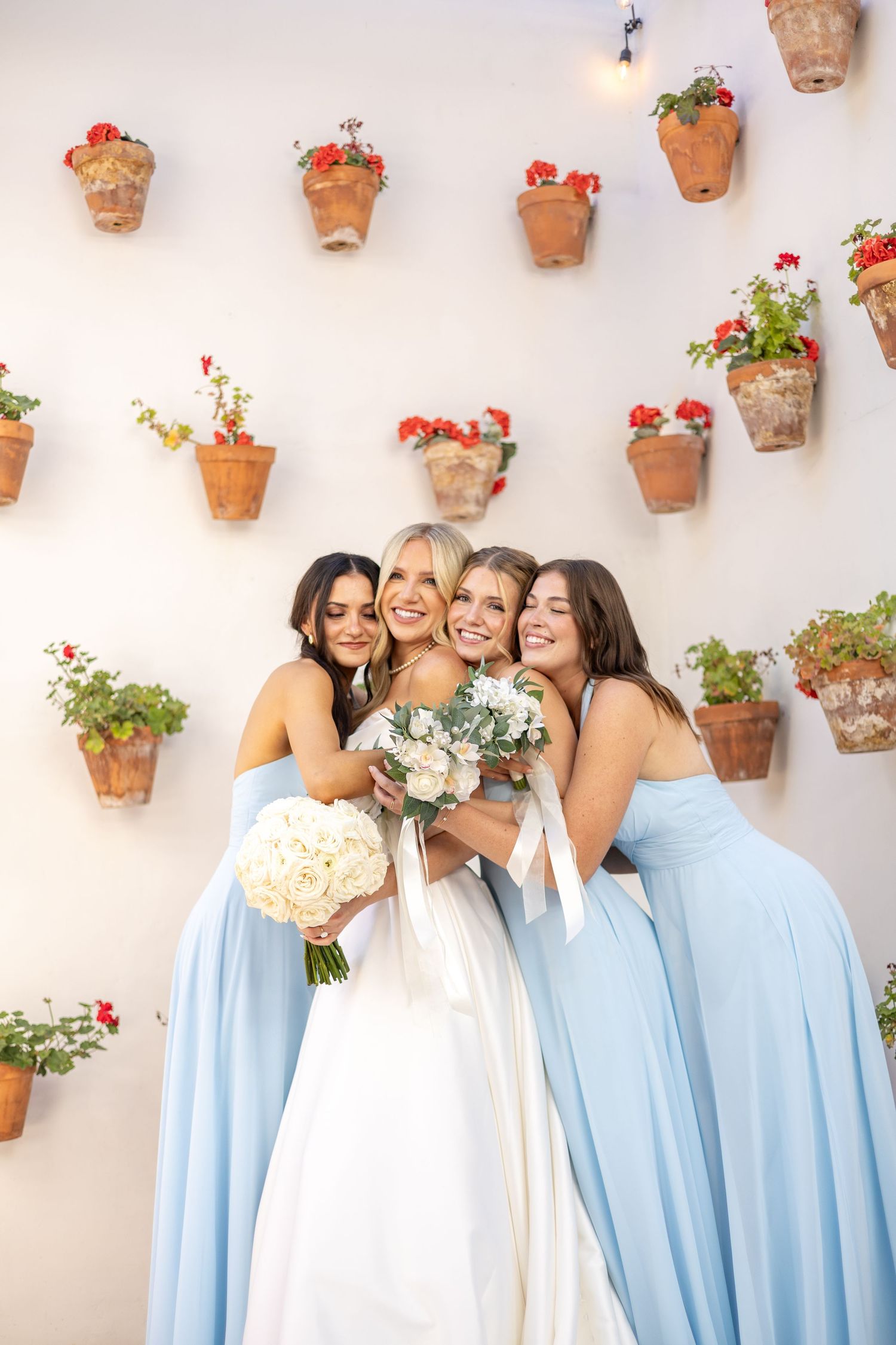Bride in white gown holds bouquet with three bridesmaids in light blue dresses against a wall with terracotta flower pots.