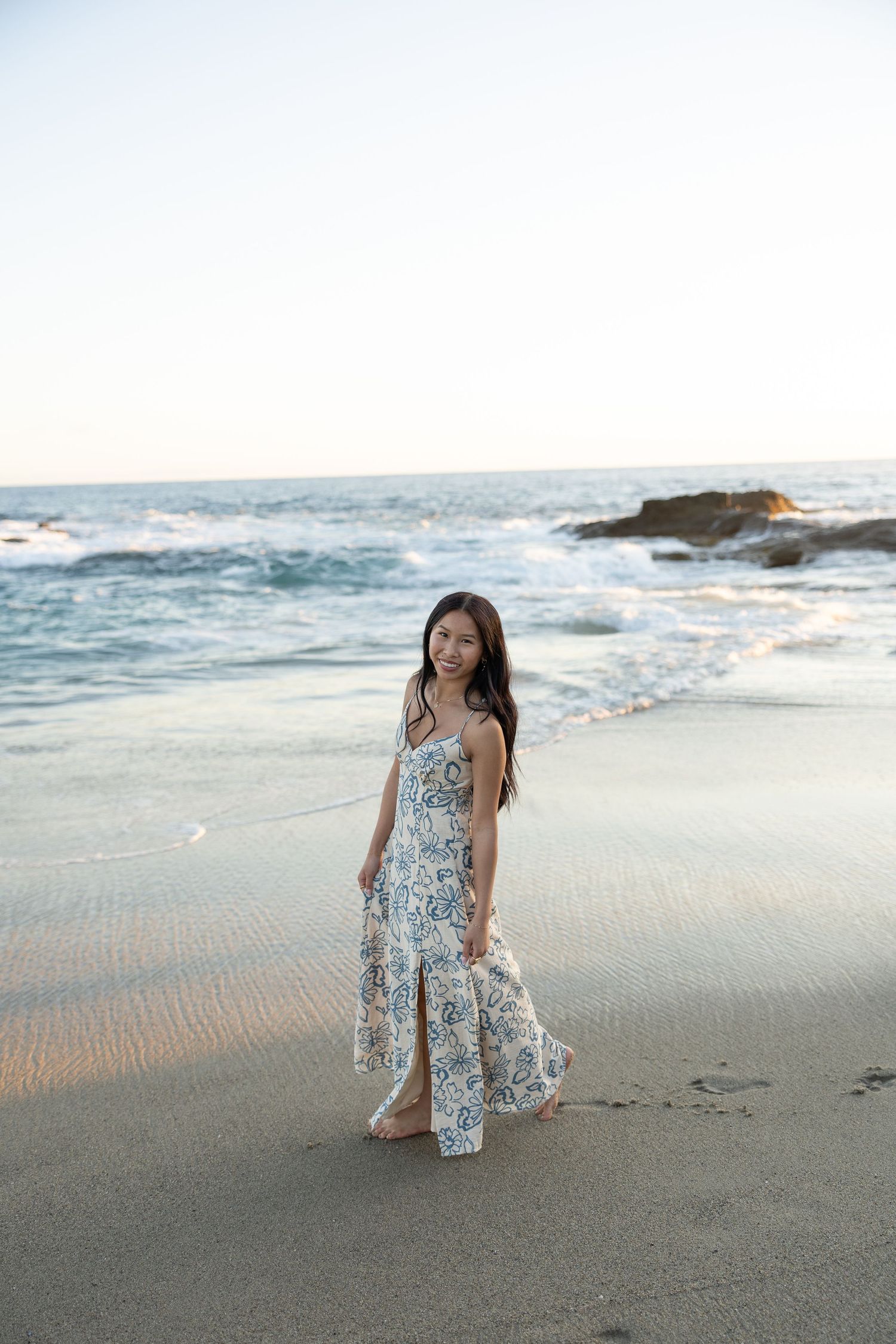 Young woman in floral maxi dress standing on sandy beach with ocean waves in background.