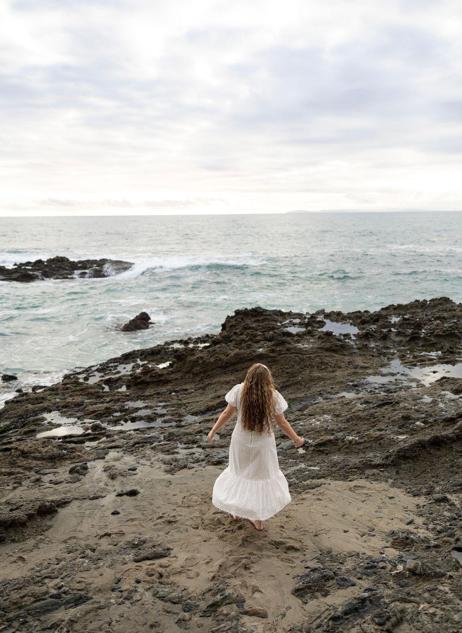 A person in a flowing white dress sits on rocky coastal cliffs overlooking turbulent ocean waves.