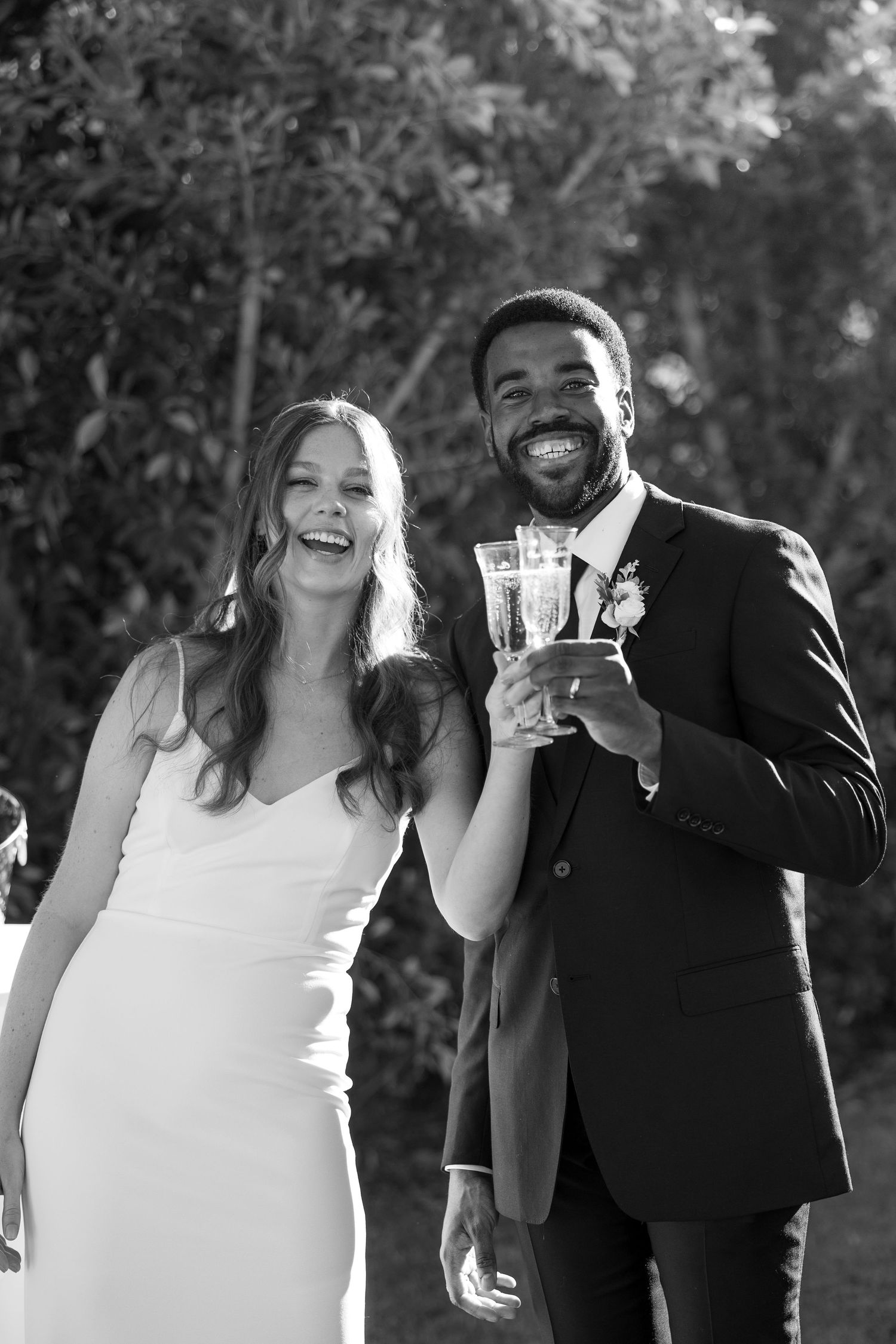 Black and white photo of a smiling bride and groom toasting with champagne glasses at their outdoor wedding.