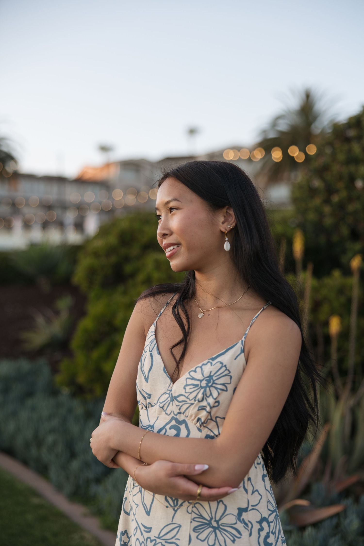 Young Asian woman in floral sundress with pearl earrings stands in garden at dusk, arms crossed, smiling softly.