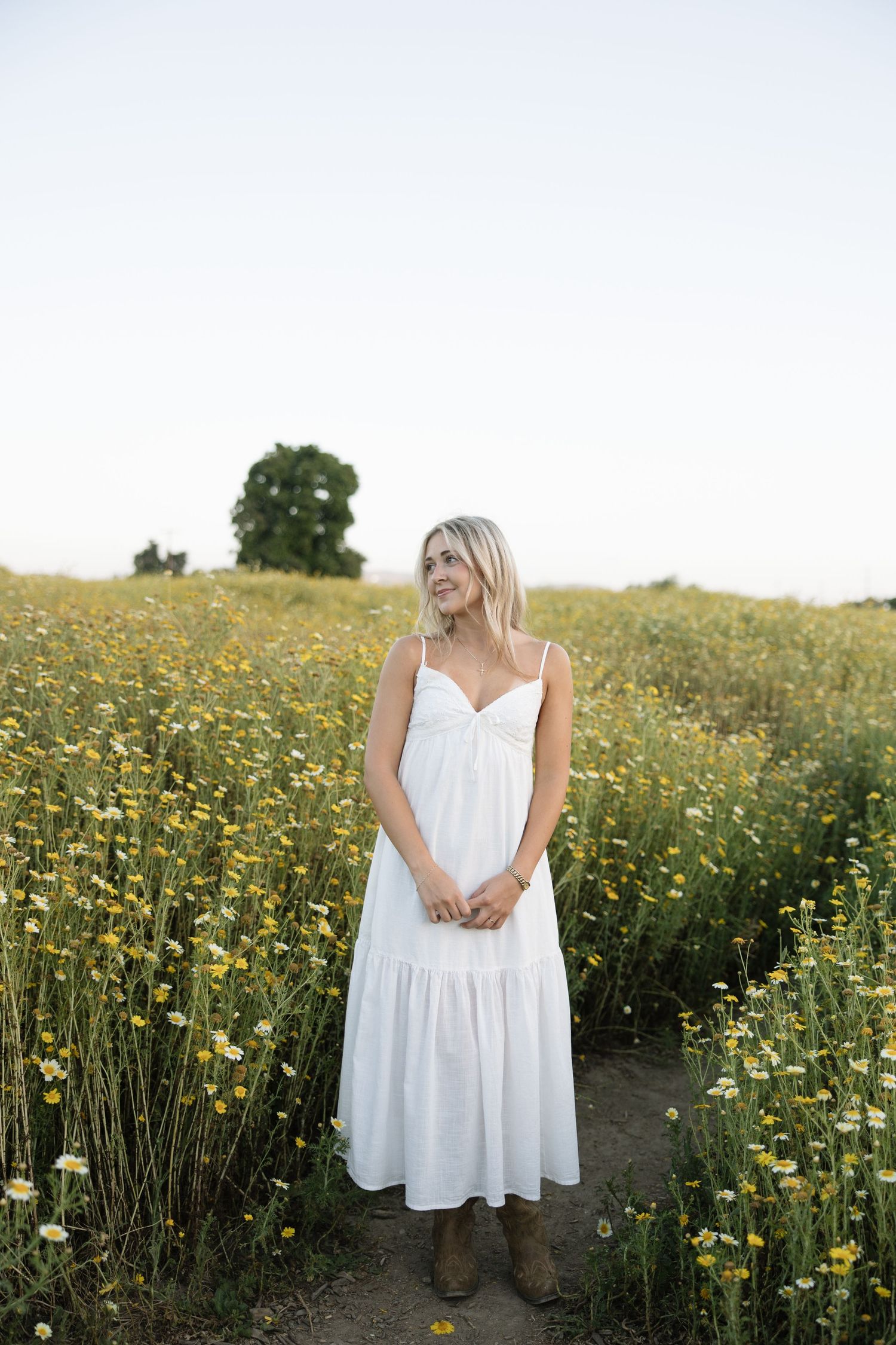 Woman in white sundress standing in a field of yellow wildflowers, smiling softly on a sunny day.