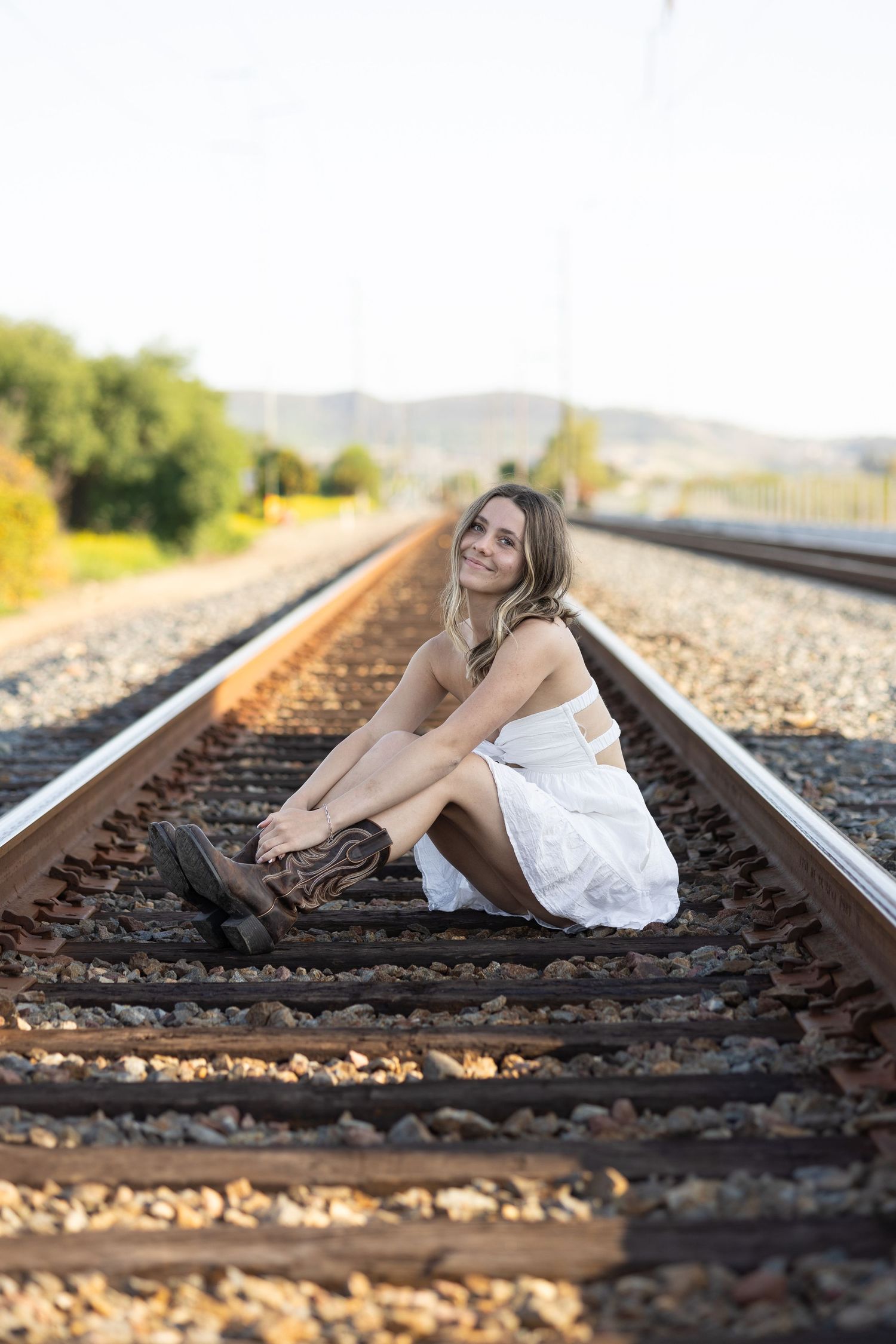 Person in white dress poses on train tracks at sunset in rural setting.