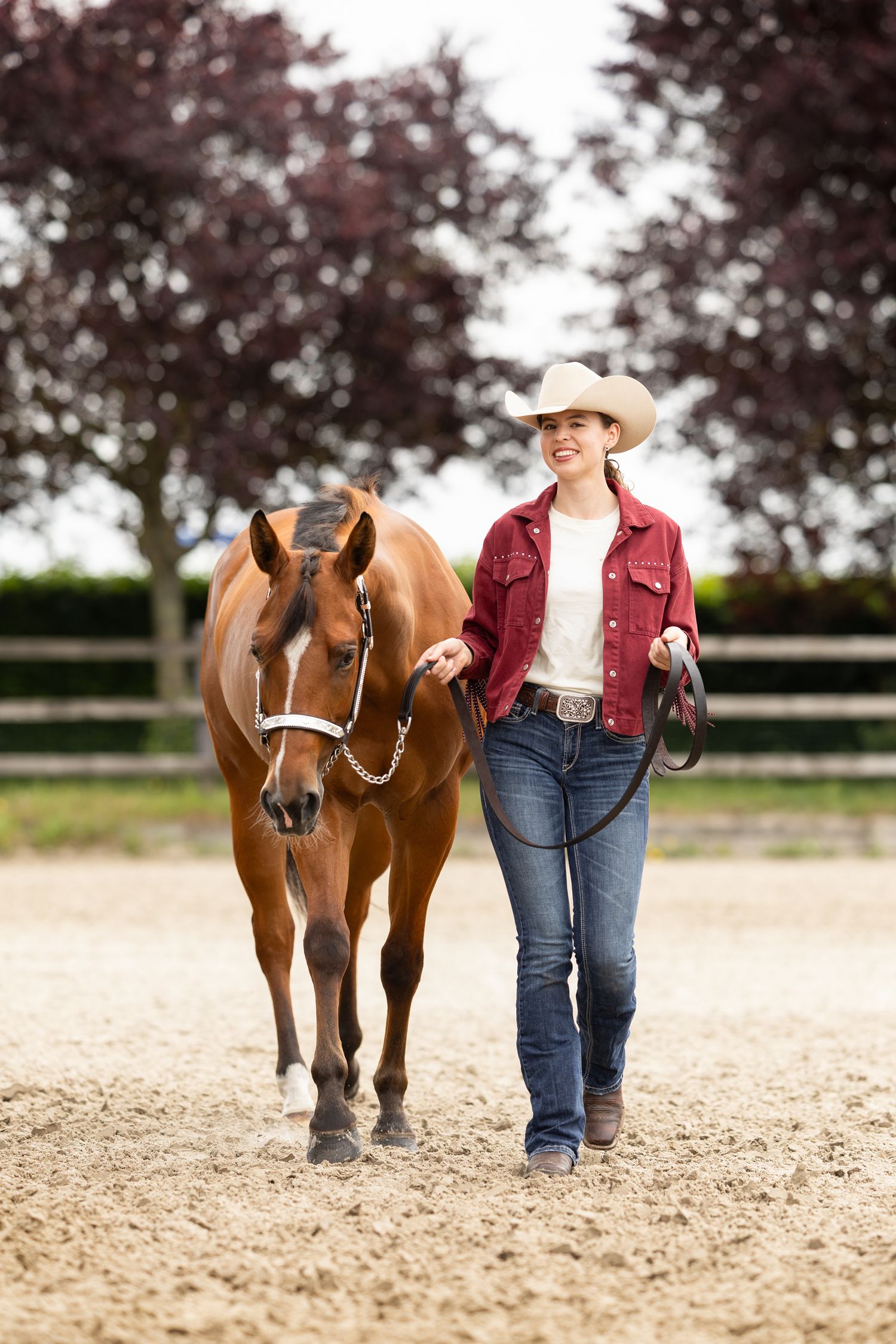 Brown horse and rider in red jacket walking together in outdoor arena with trees in background.