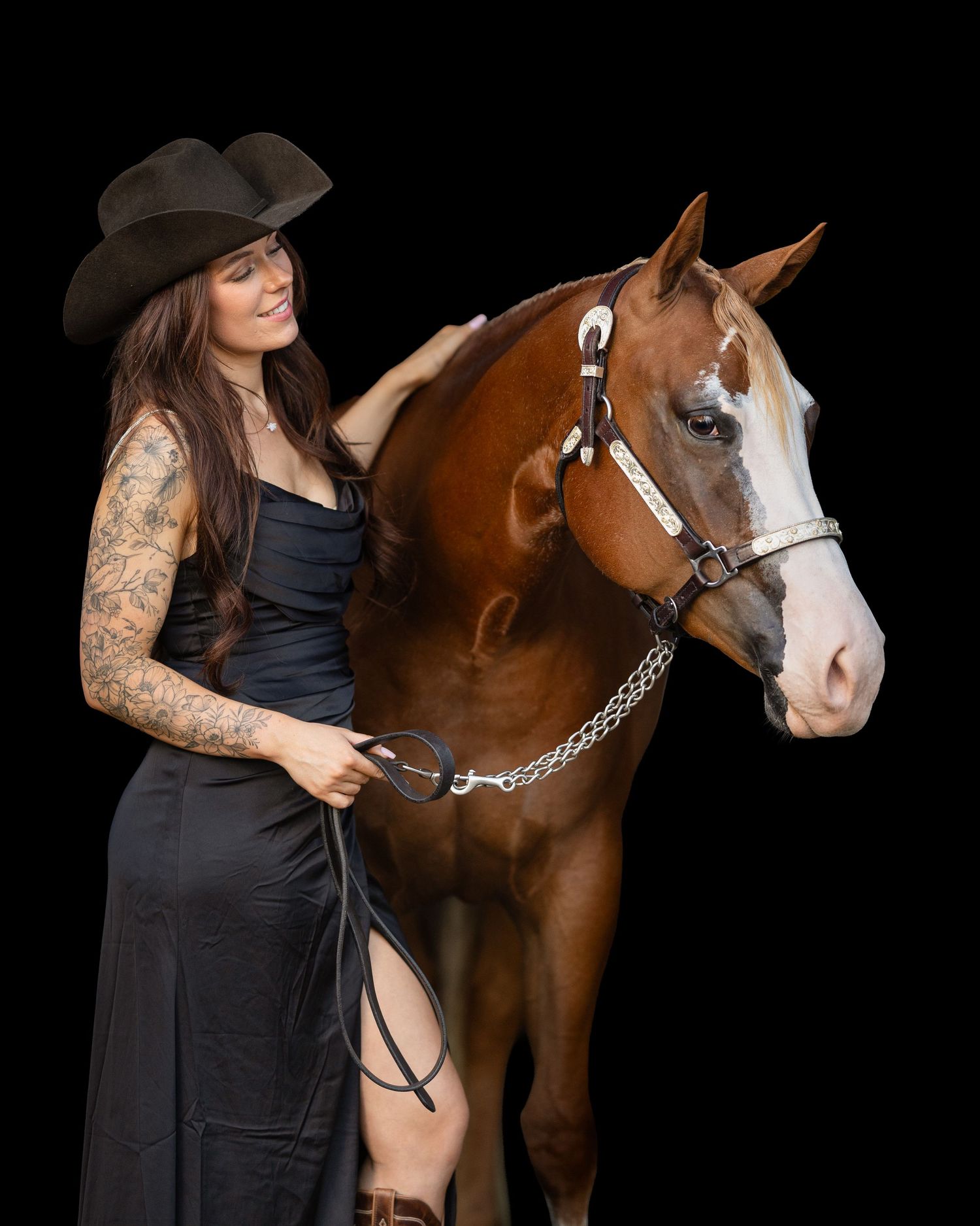 Tattooed woman in black dress and cowboy hat posing with a chestnut horse on dark background.
