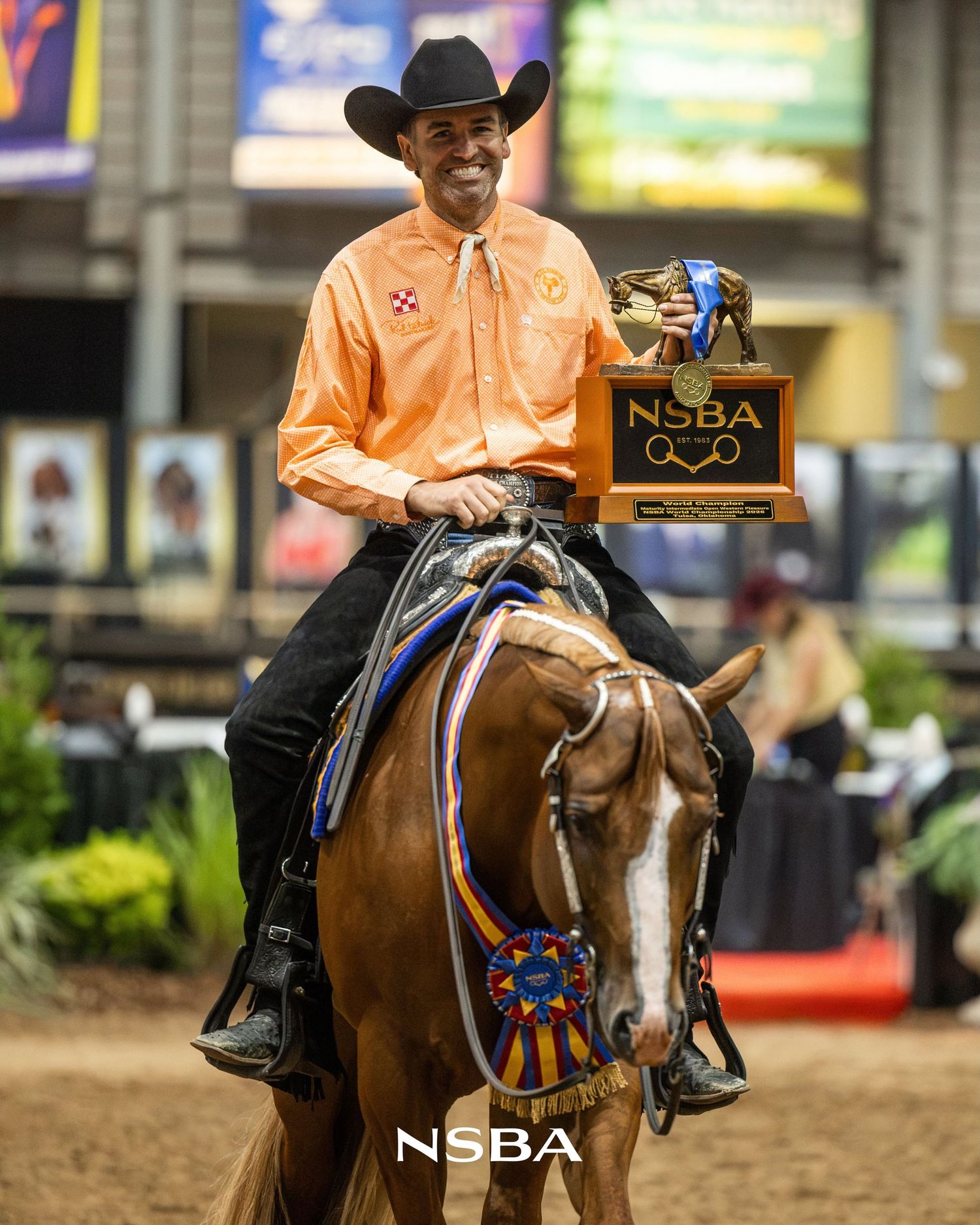 Smiling Anthony Montes in orange shirt on chestnut horse holds NSBA trophy with blue ribbons at the NSBA World Show