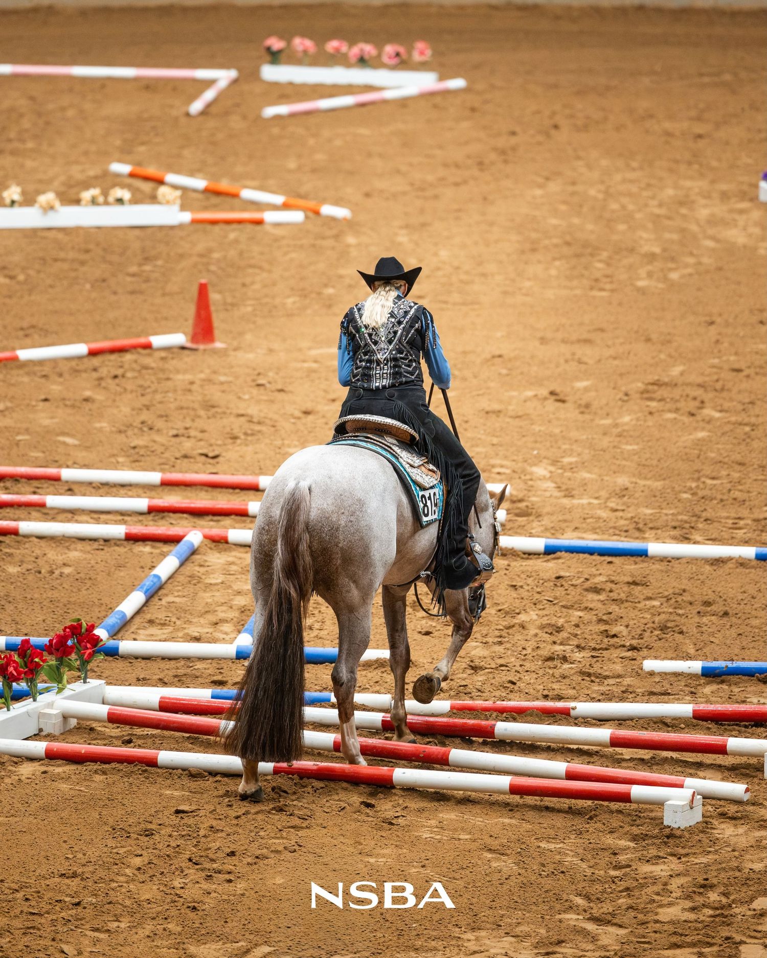 Rider in black hat navigates a gray horse through poles at an NSBA equestrian competition on a dirt arena.