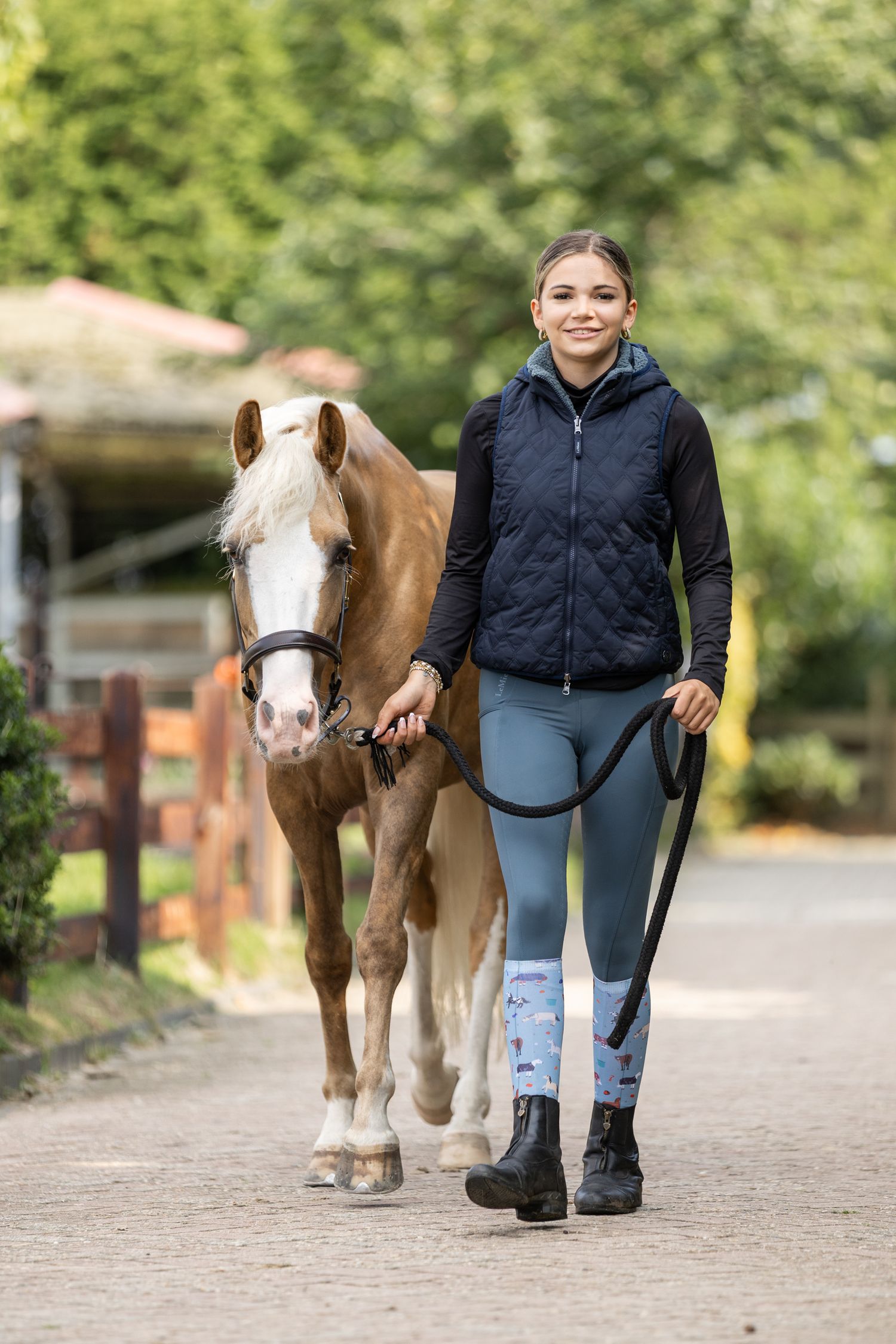 Brown and white pony with equestrian handler walking on a sandy path surrounded by greenery.