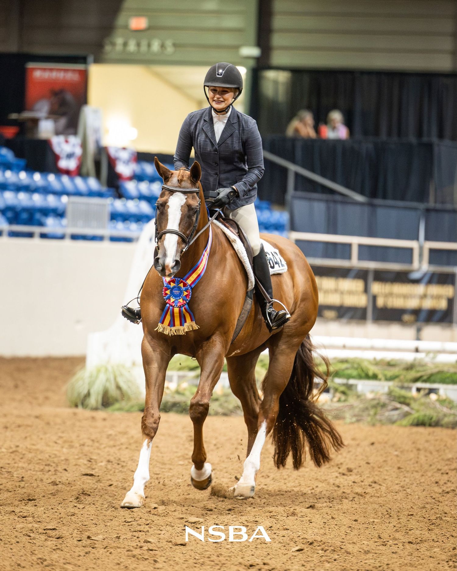 A smiling equestrian rider on a chestnut horse displaying a blue ribbon award at an the NSBA World Show