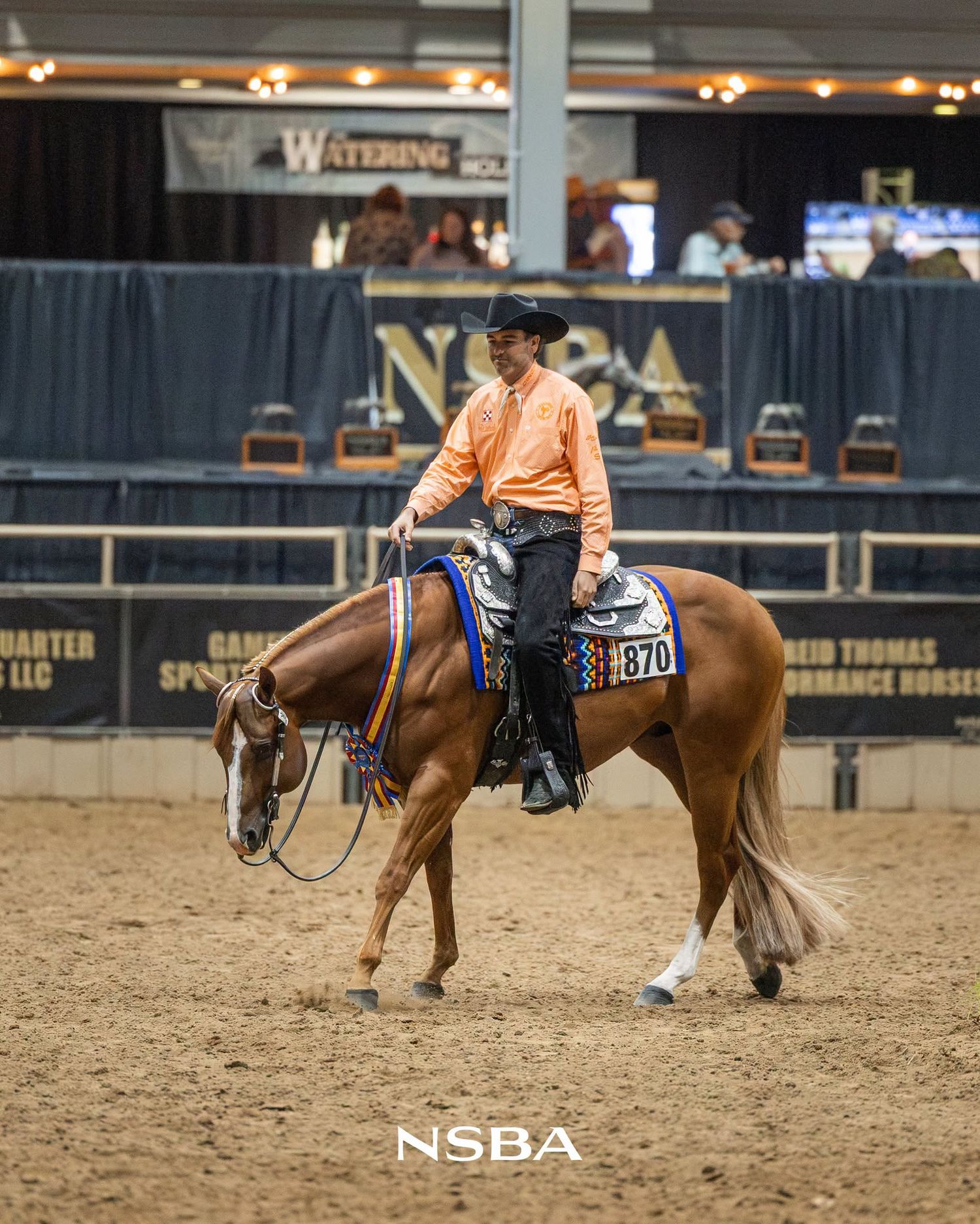 Rider in orange shirt on chestnut horse competing at NSBA equestrian show in indoor arena.