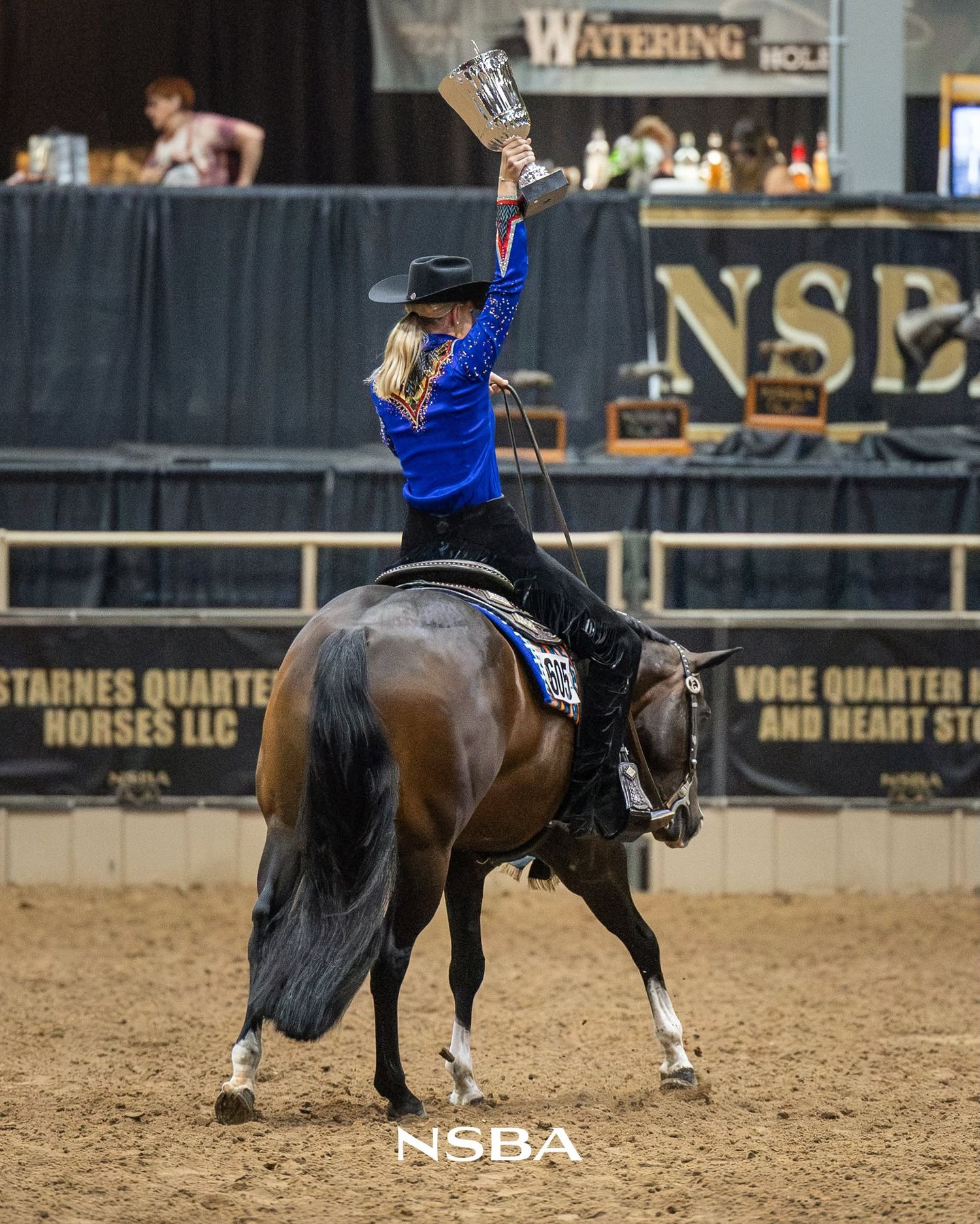 Female equestrian rider celebrates victory on horseback at NSBA horse show, raising trophy overhead.