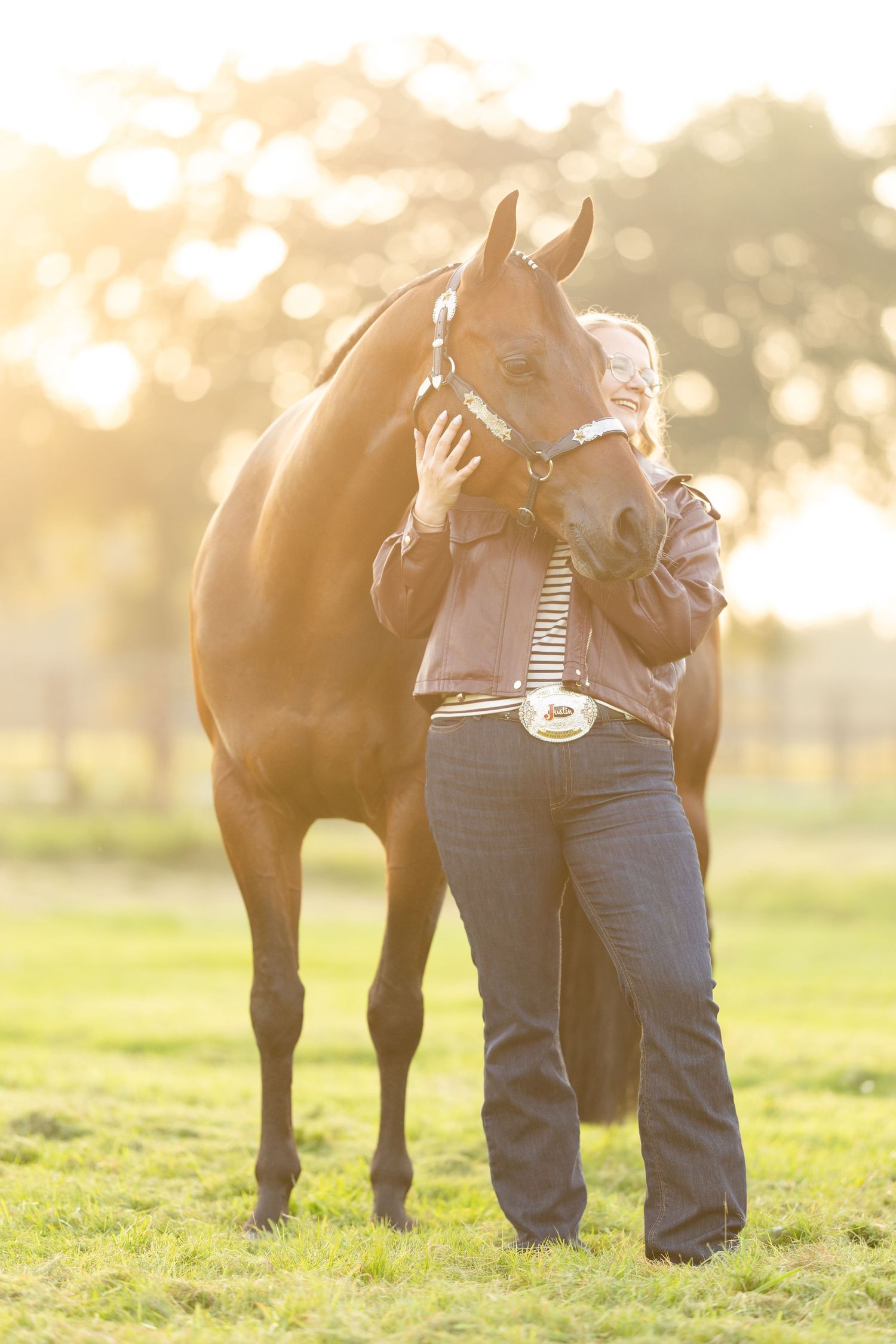 A woman affectionately embraces a dark bay horse in a sunlit green field with warm golden bokeh background.