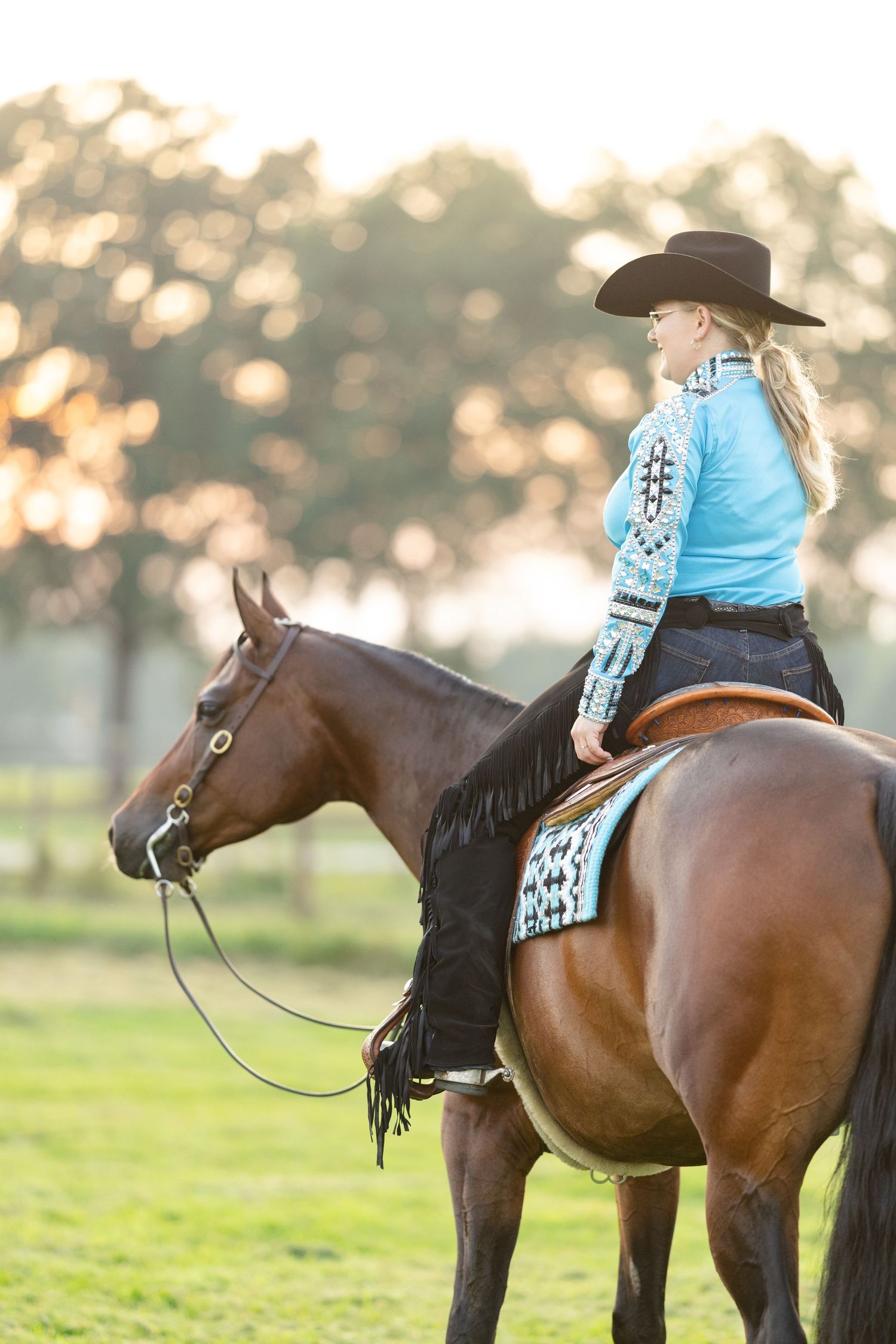 A blonde woman in a turquoise western shirt and black cowboy hat rides a chestnut horse at sunset in a green field.