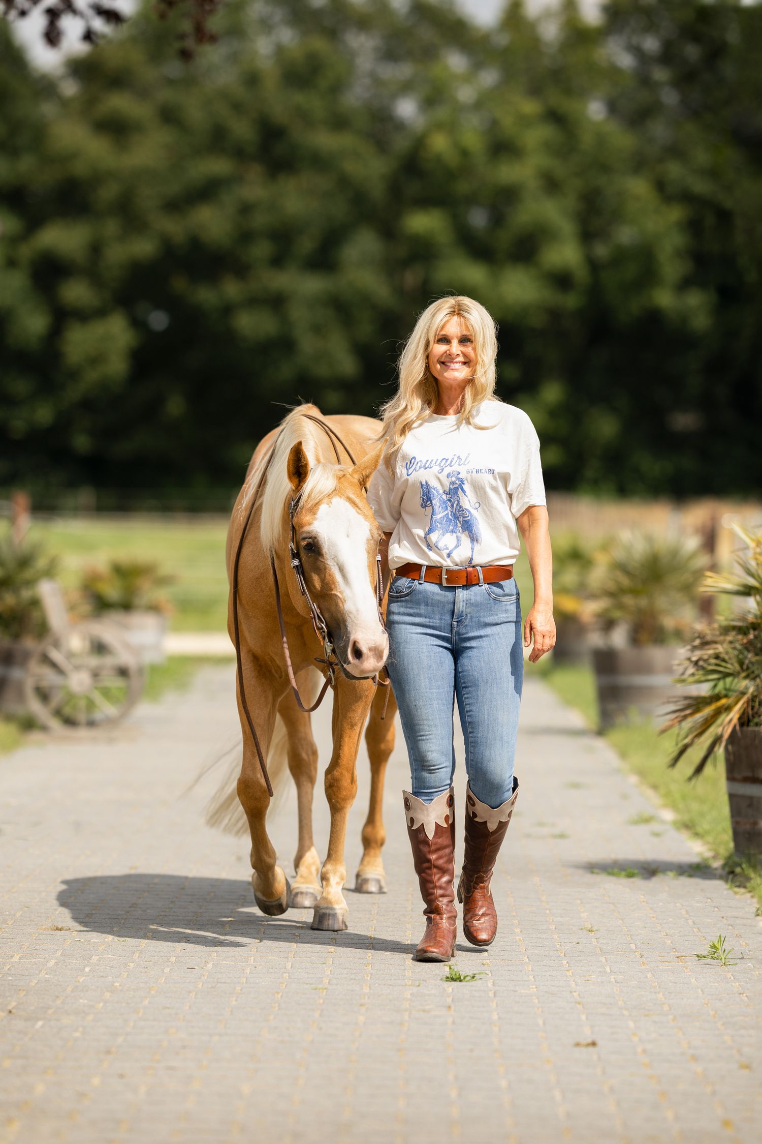 Woman in western attire walking with a palomino horse on a path surrounded by greenery.