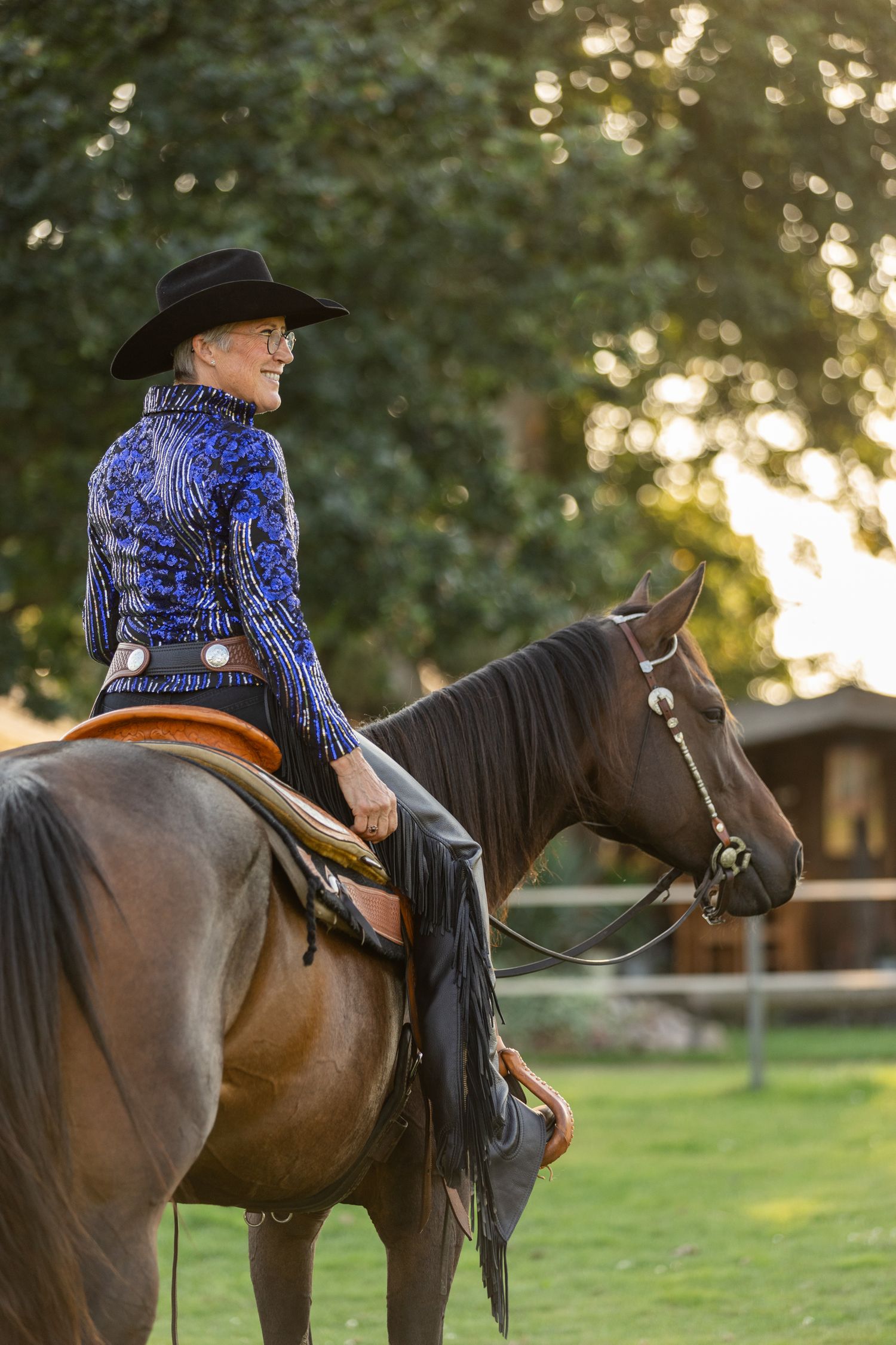 Equestrian rider in blue western show shirt and black hat sitting astride a brown horse at sunset.
