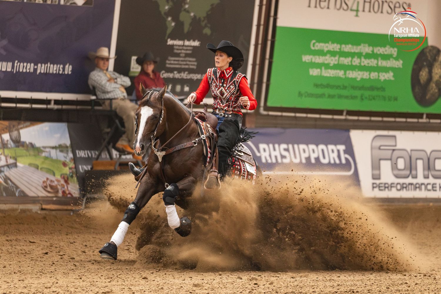 A reining horse and rider perform a sliding stop in an indoor arena, kicking up sand during a competition.
