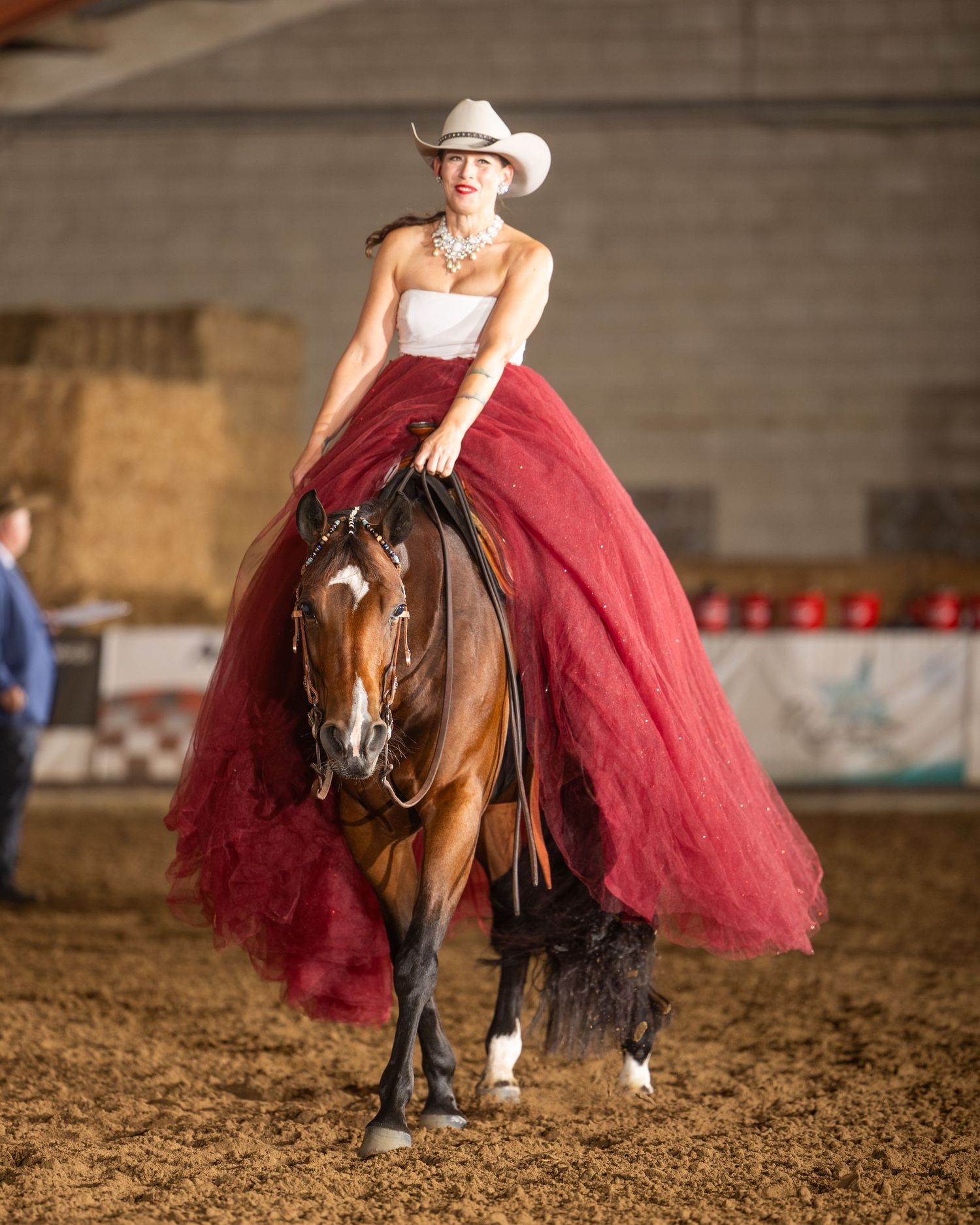 Woman in red ballgown and cowboy hat riding a bay horse in an indoor arena during an equestrian show.